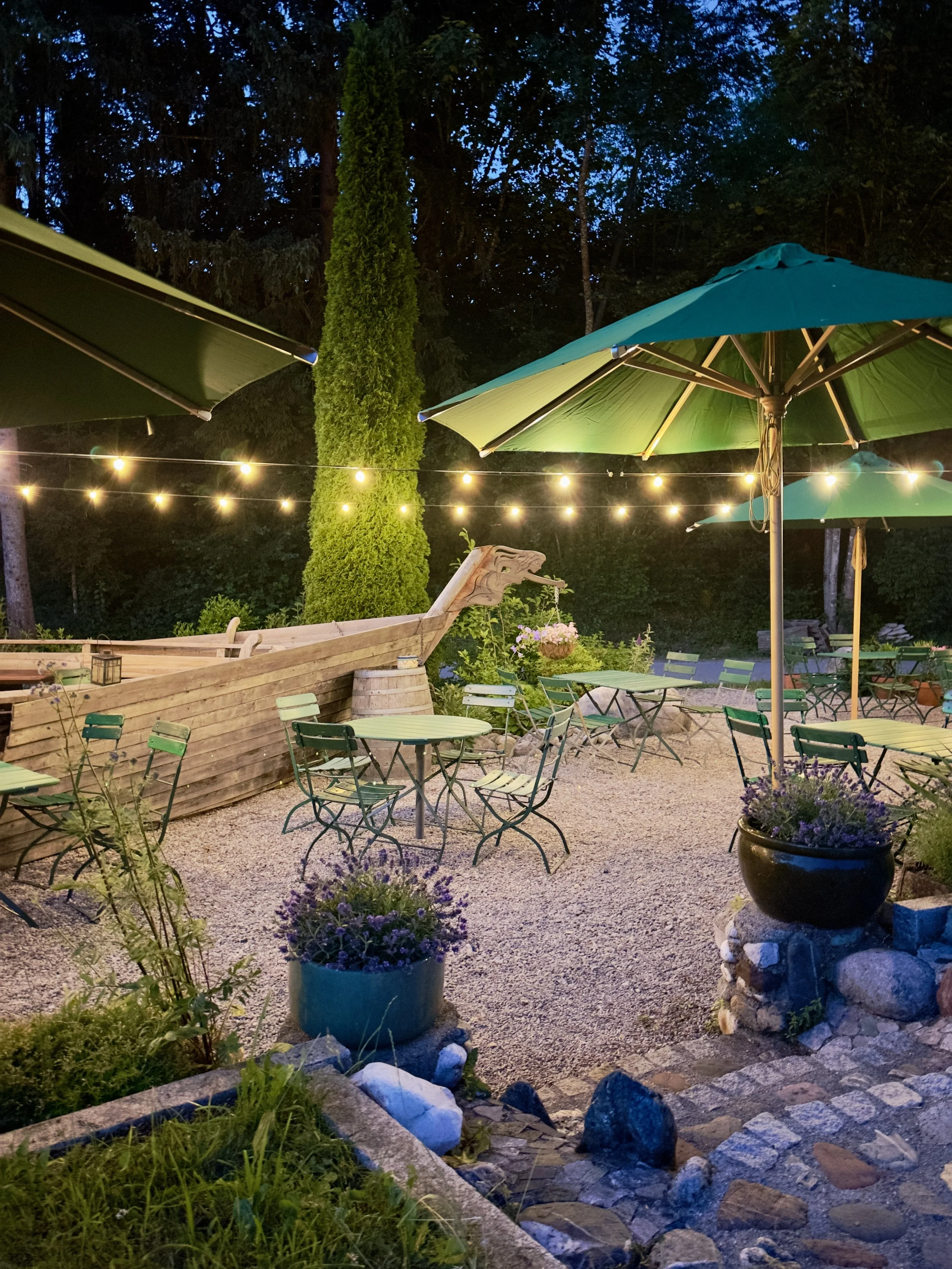 Ein gemütlicher Außenbereich eines Cafés oder Restaurants bei Nacht, mit Tischen, Stühlen, Sonnenschirmen, Lichterketten, Pflanzen und einer mittelalterlichen Drachenboot-Dekoration.
