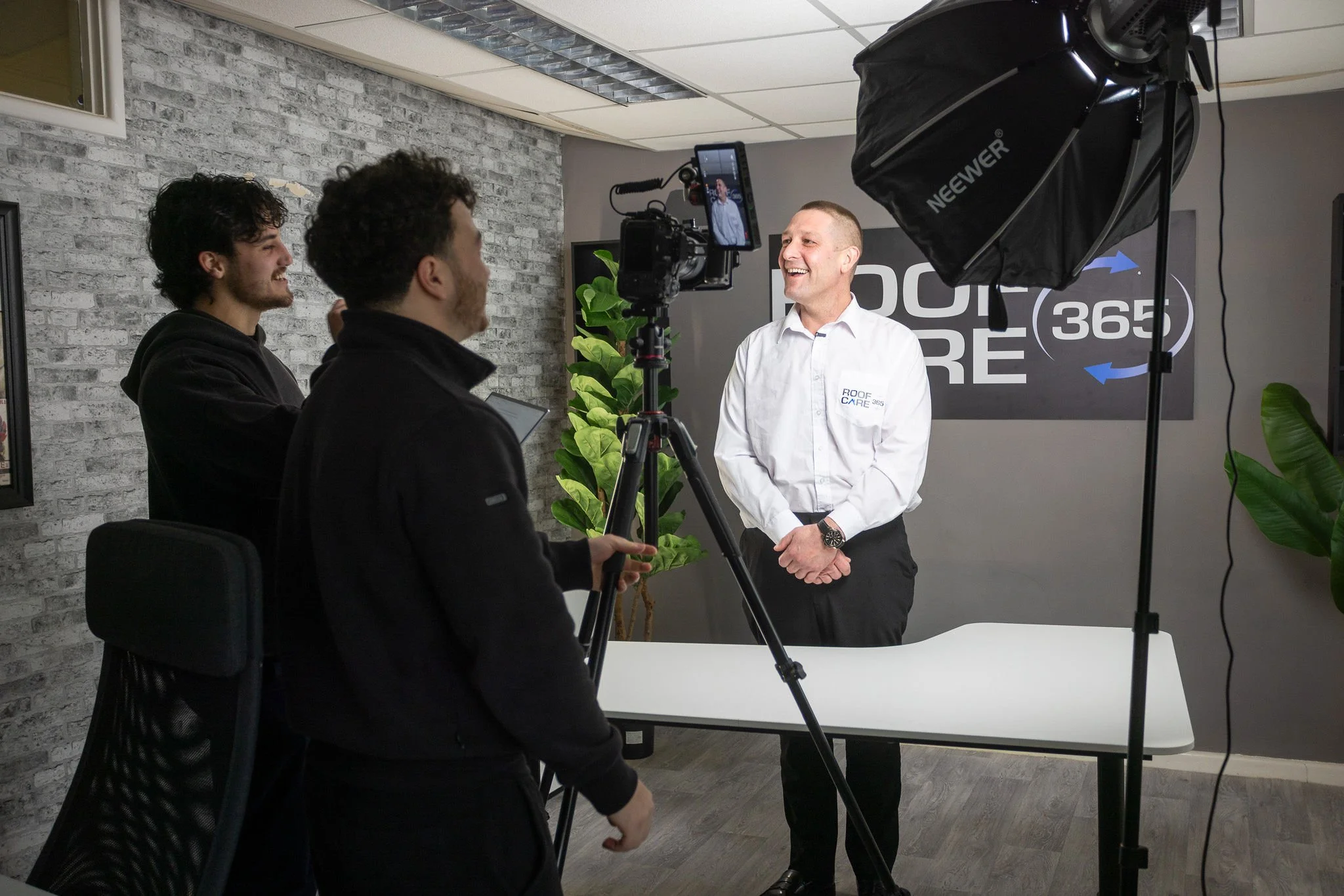 A video recording of a man in a white shirt with 'Roof Care' embroidered on it, smiling and standing in front of a camera. Two young men are operating the camera and equipment in a professional recording studio with a gray brick wall, plants, and a logo in the background.