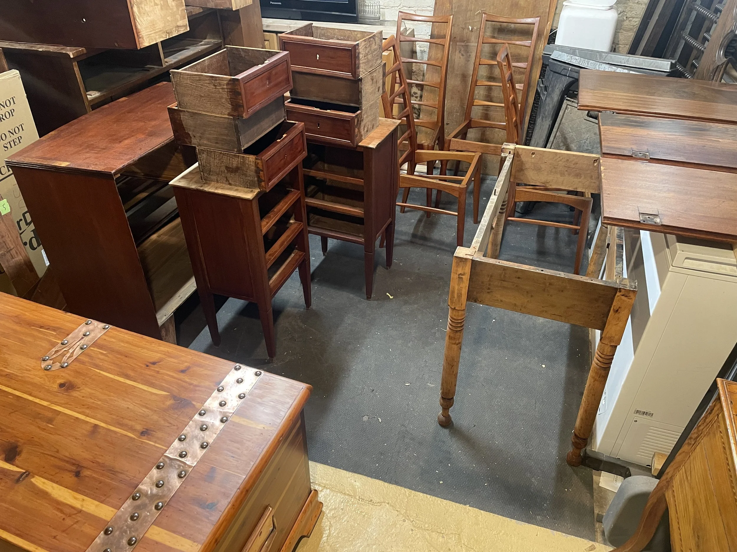 A collection of wooden furniture including chairs, tables, and shelves, arranged haphazardly in an indoor space, possibly a secondhand store or warehouse.