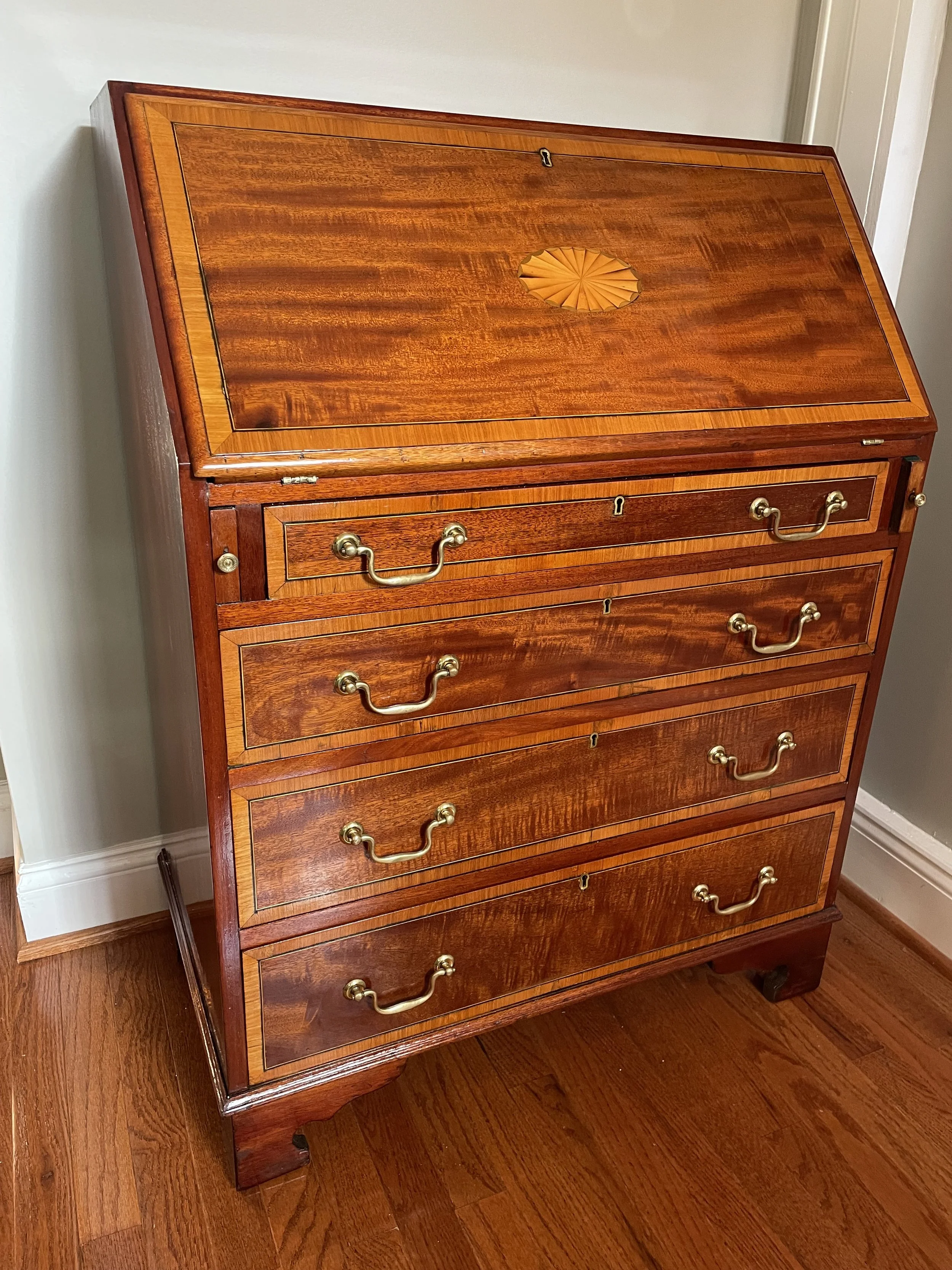 A wooden secretary desk with a slanted drop-down writing surface, brass handles, and five drawers, standing on a hardwood floor against a light-colored wall.