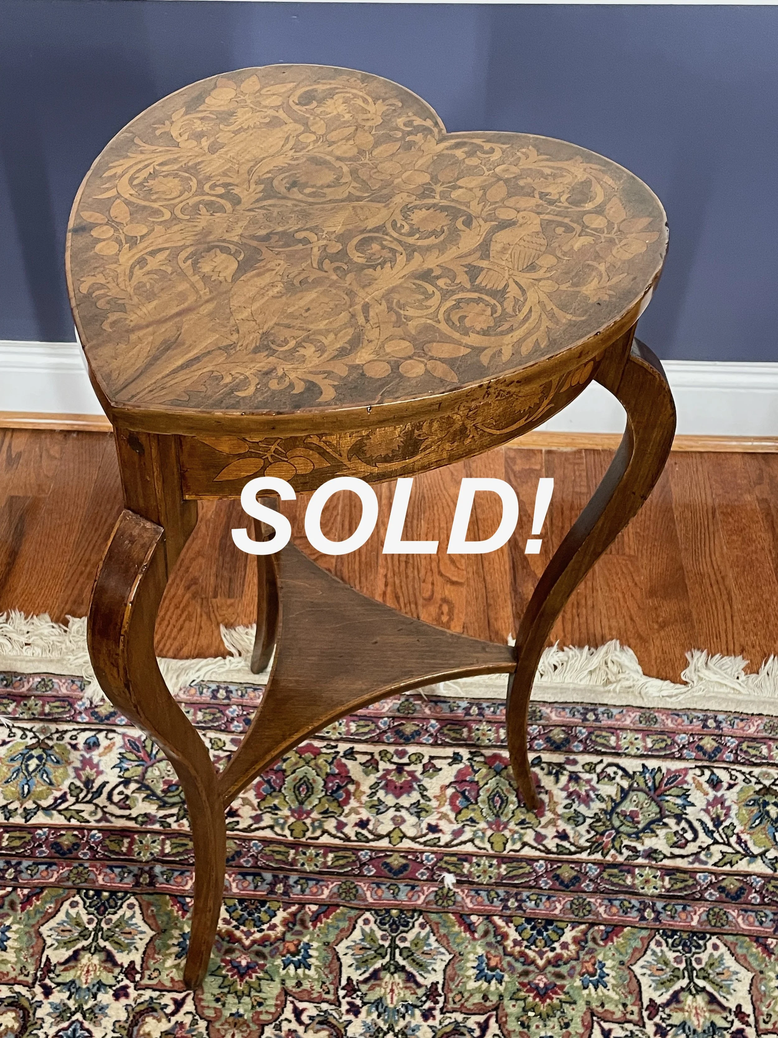 A wooden, heart-shaped table with decorated, intricate carvings on the top and curved legs, placed on a patterned rug against a blue wall. The word 'SOLD!' is overlaid on the table.