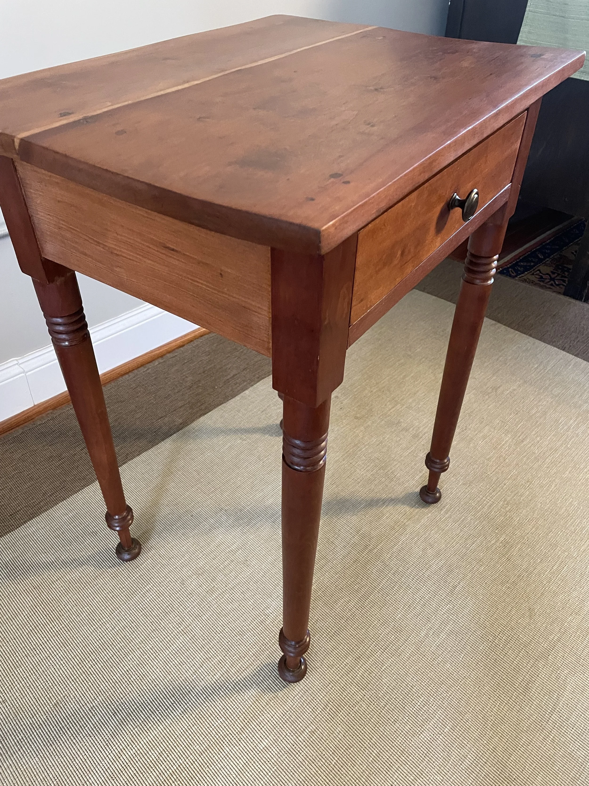 A wooden side table with a single drawer and a round metal handle, standing on four turned legs, placed on a beige rug in a room.