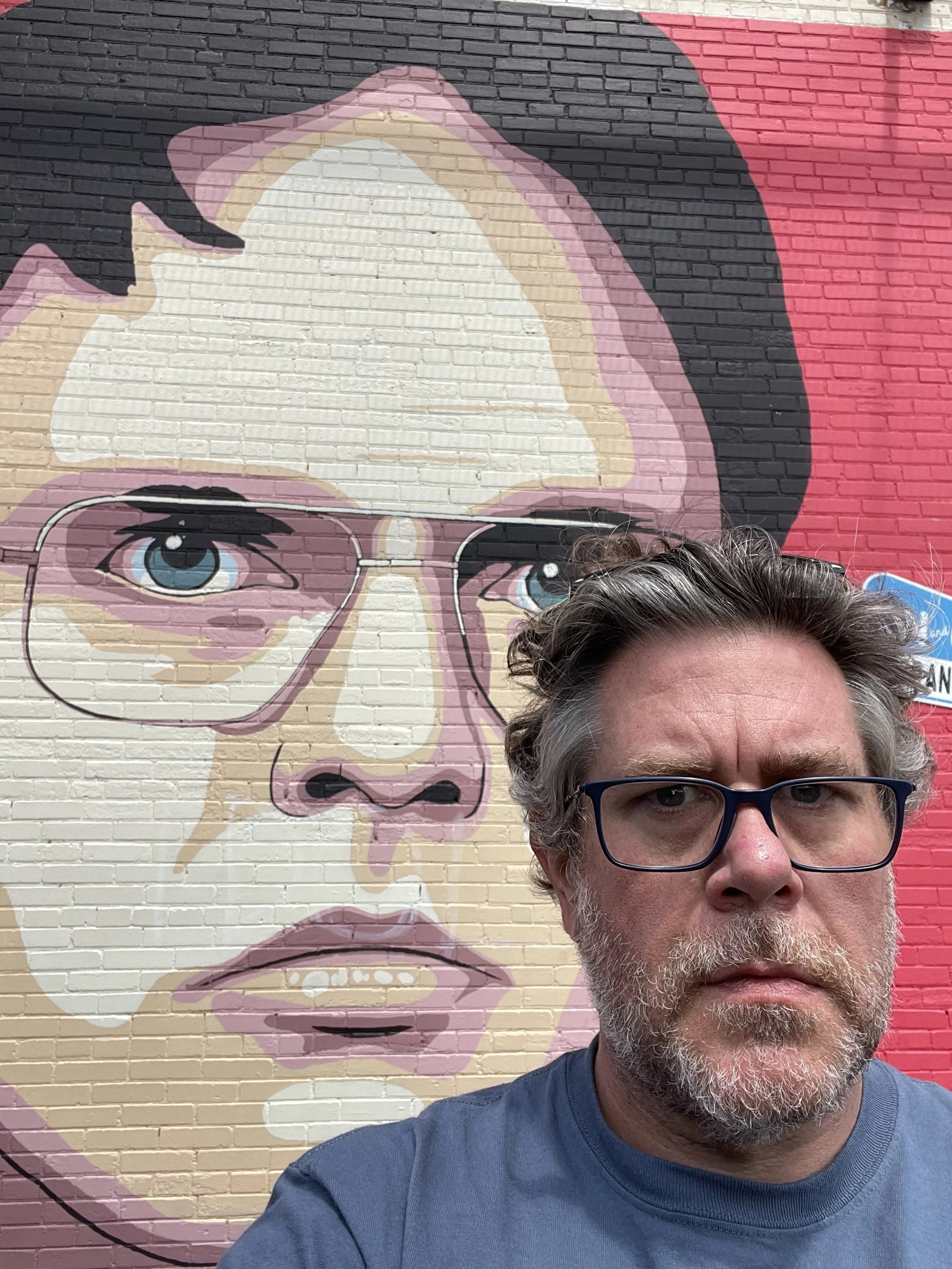 A man with glasses and gray hair taking a selfie in front of a mural of a woman with glasses and blue eyes painted on a brick wall.
