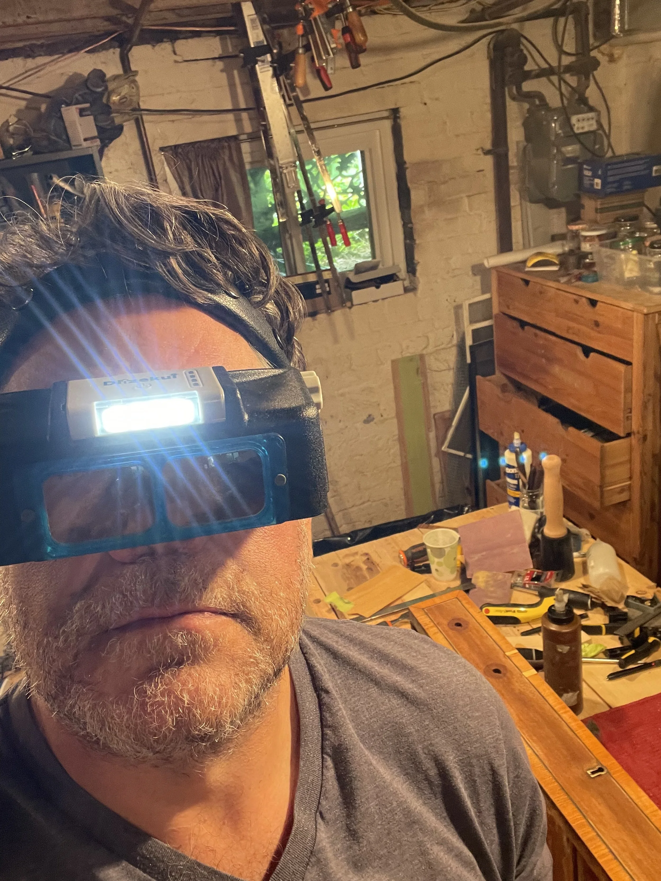 A man wearing a headlamp in a cluttered workshop with tools, wood, and a window showing greenery outside.