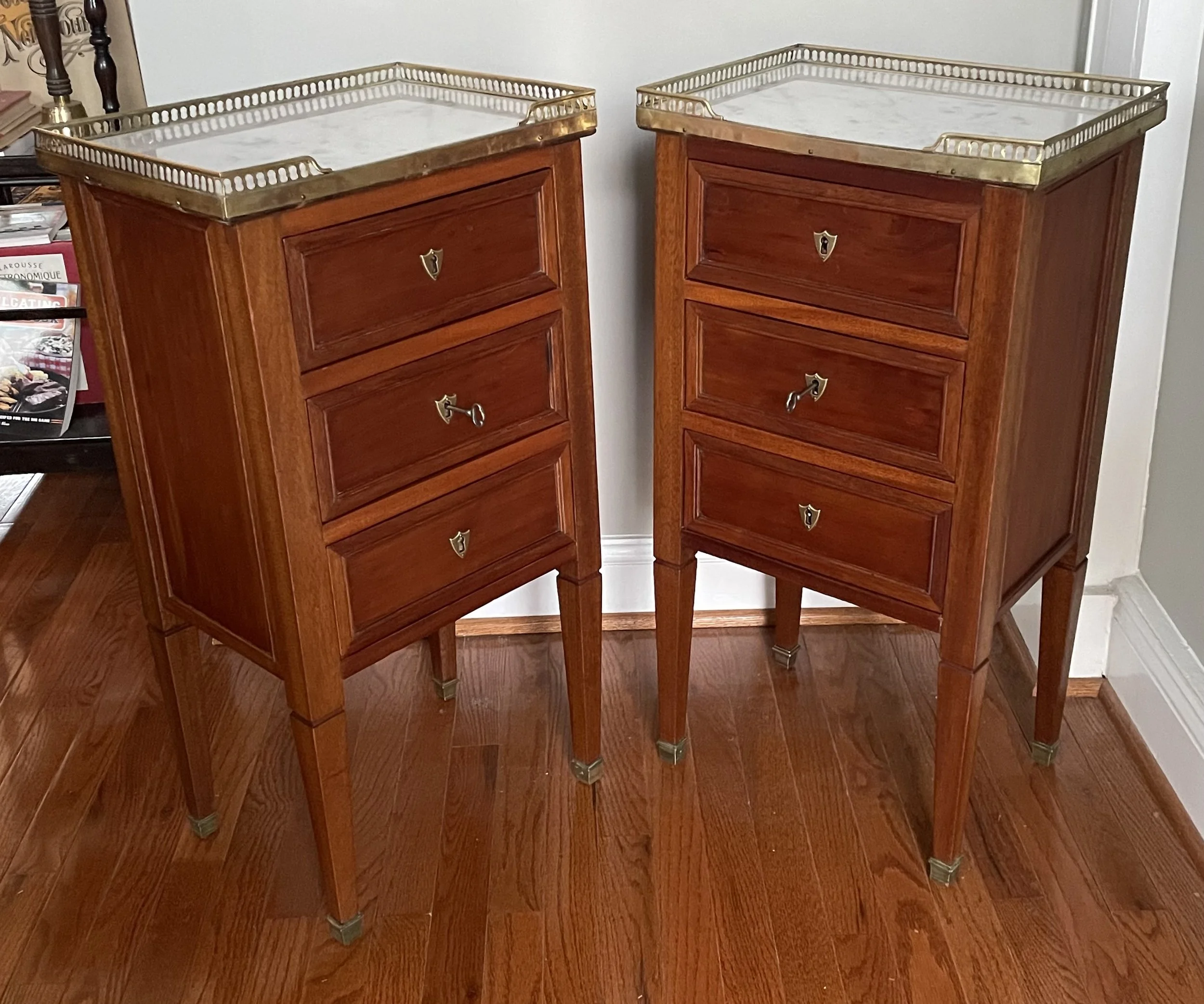 Two vintage wooden bedside tables with four drawers and brass accents, topped with brass trays, on a hardwood floor in a room.