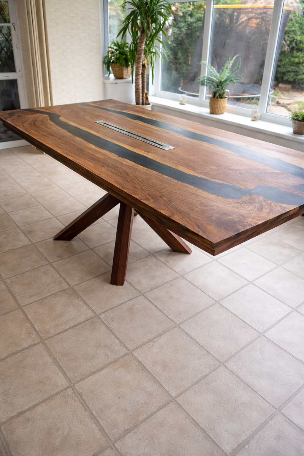 A wooden dining table with black resin inlays and a built-in power outlet on top, positioned on a tiled floor in front of windows with potted plants outside.