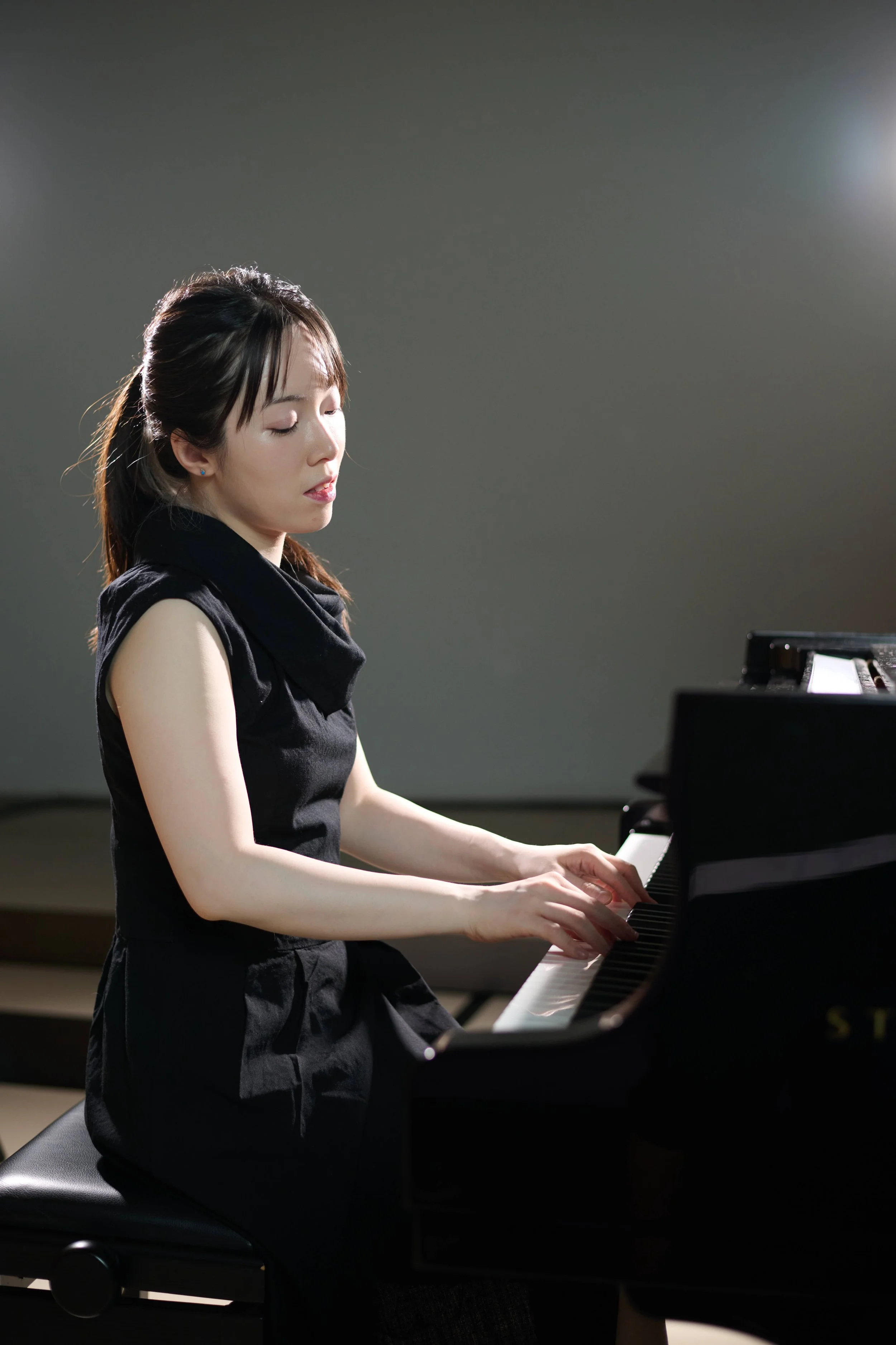 Jessica Wu, a young woman, playing a grand piano with a focused expression, dressed in black, in a music studio or performance space.