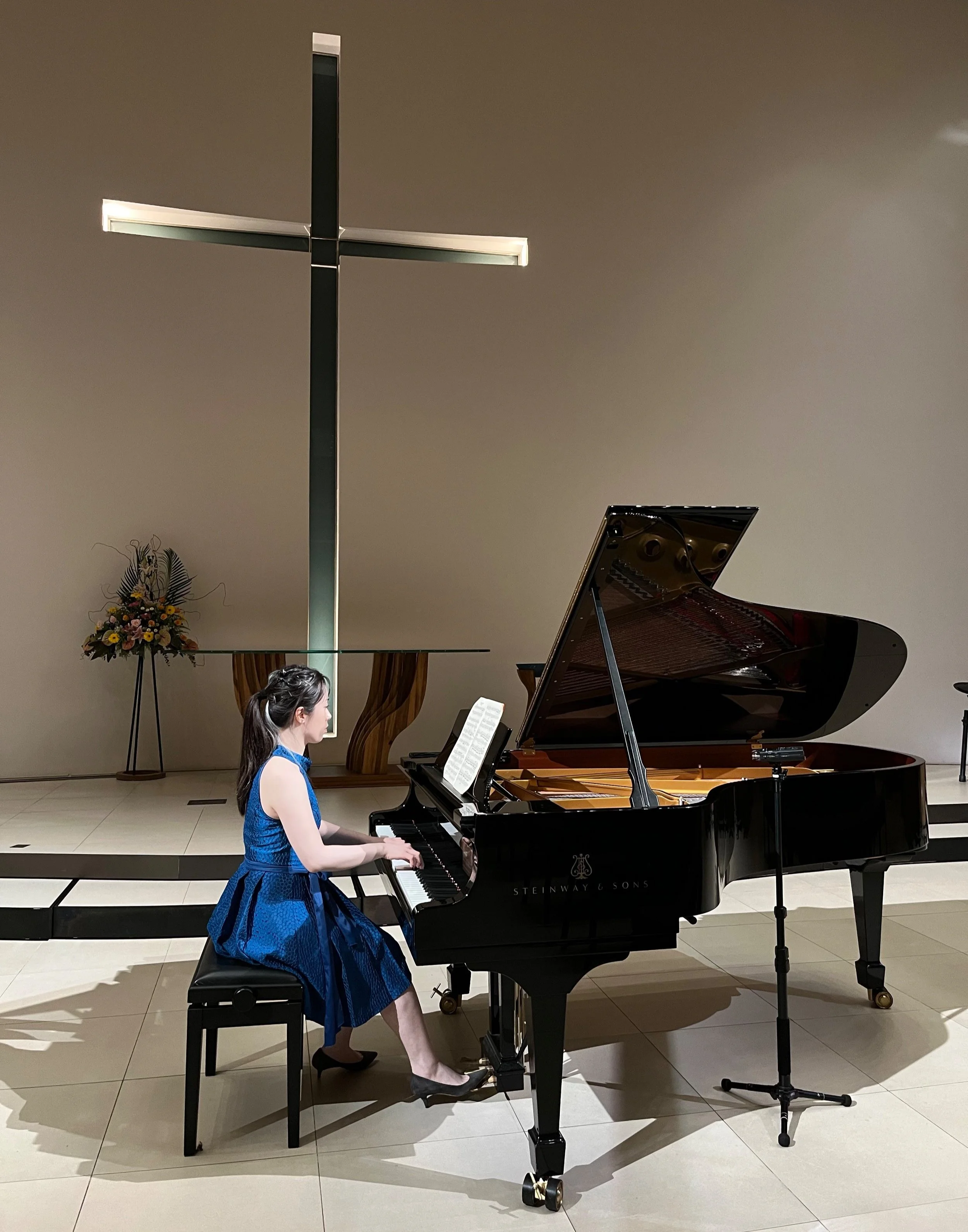 Jessica Wu, a young woman, in a blue dress playing a grand piano in a church with a large cross on the wall behind her.