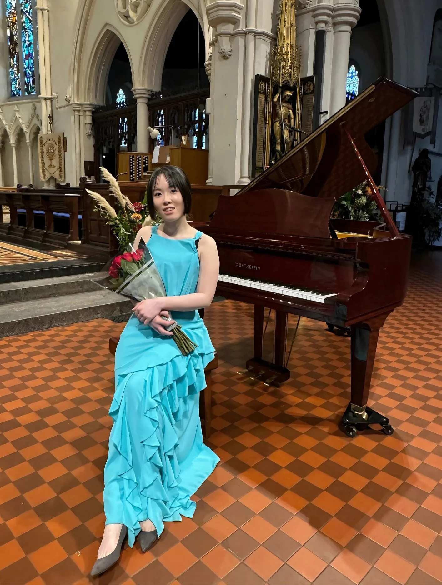 Jessica Wu, a young woman, in a blue dress holding a bouquet of flowers, sitting beside a grand piano inside a church with high arched ceilings and stained glass windows.