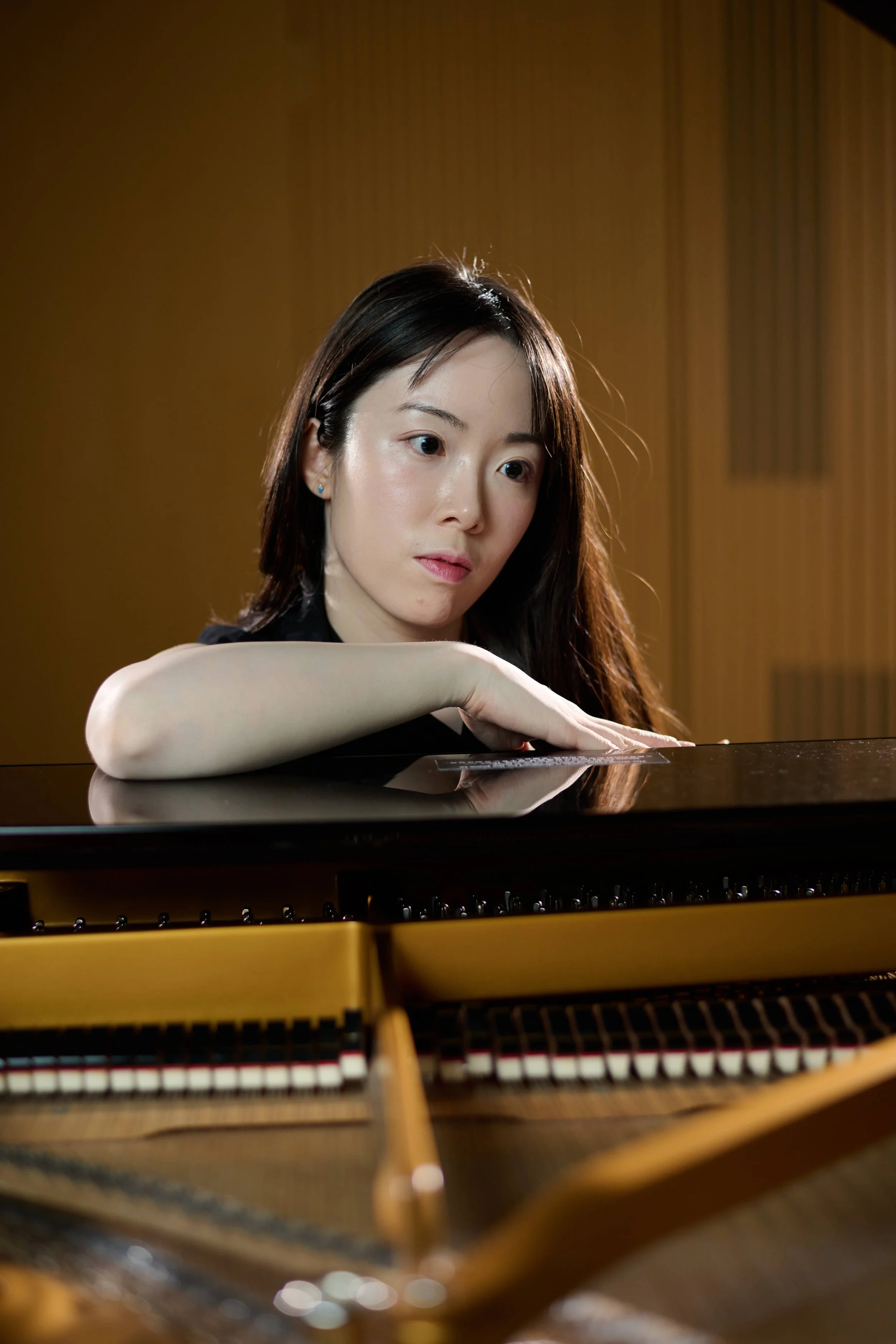 Jessica Wu, a young woman, with dark hair leaning on the piano, looking down at the keys, with an interior wooden acoustic panel background.
