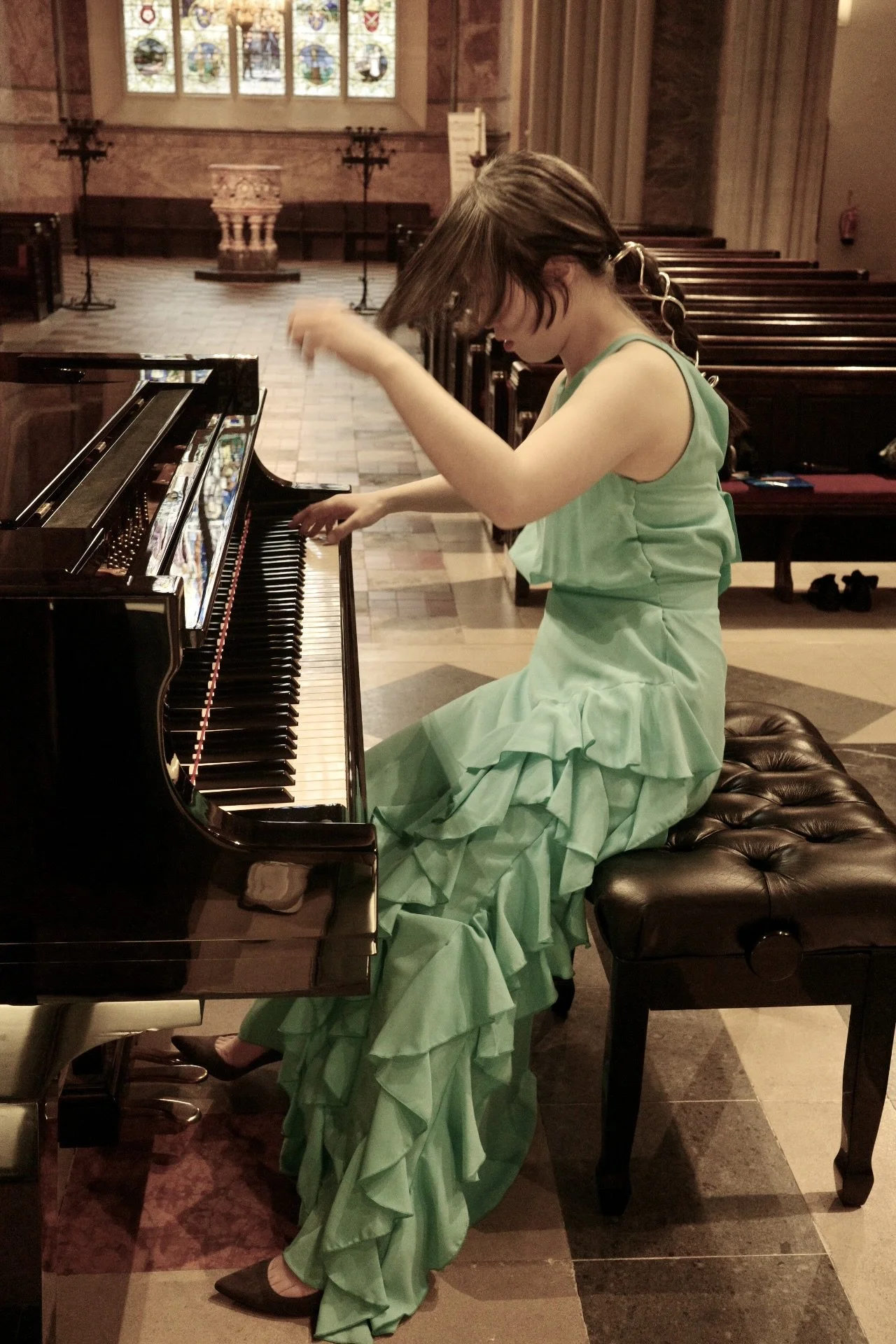 Jessica Wu, a young woman, wearing a light green dress with ruffles, sitting on a black piano bench, playing a black grand piano inside a church with stained glass windows.