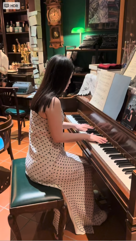 Jessica Wu, a young woman, with long dark hair playing a piano in a cozy, vintage-style bar with shelves, clocks, and various decorative items.