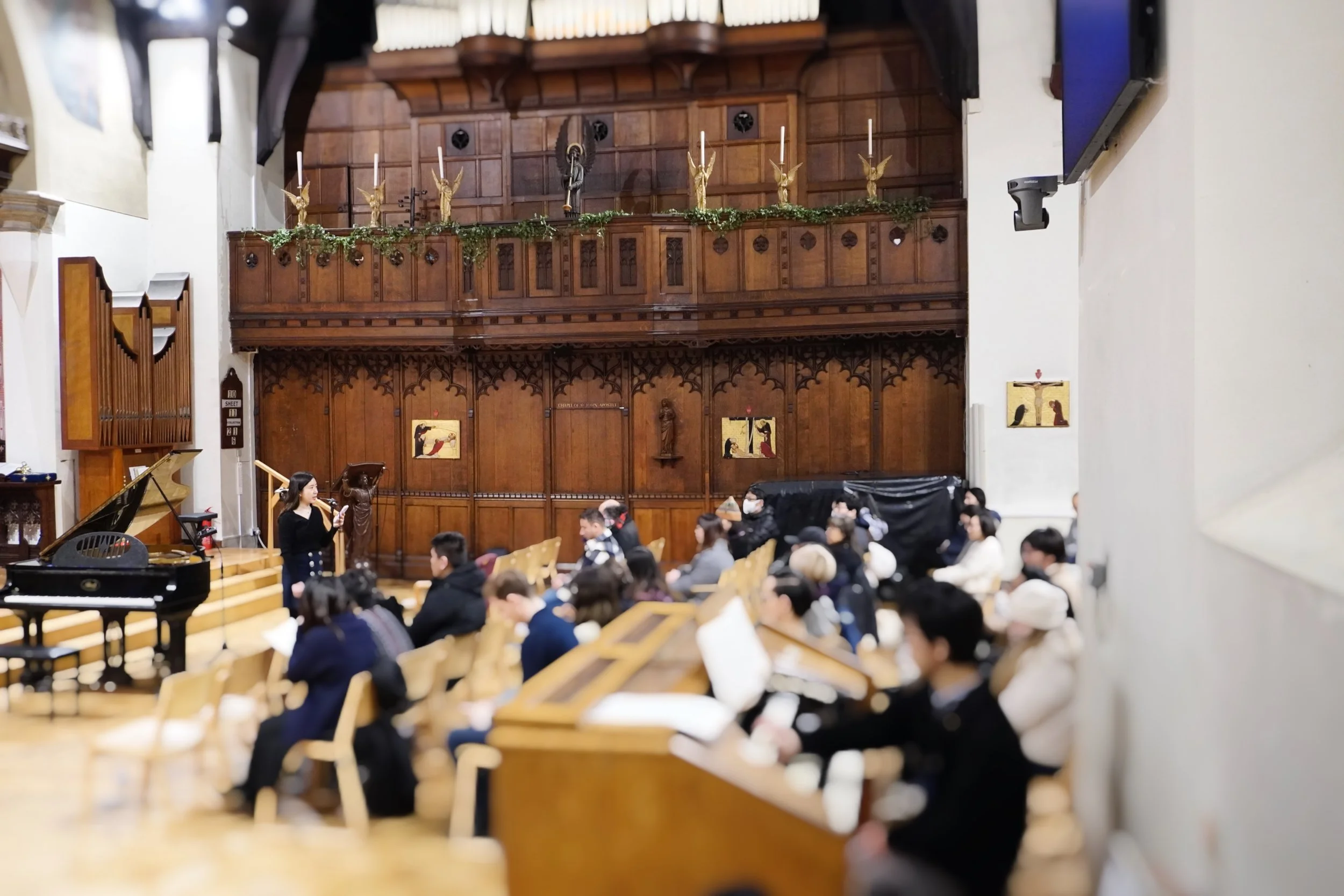 A musical performance inside a church or concert hall with a grand piano and a woman singing into a microphone. Audience members sit on wooden chairs, some holding papers, watching the performance. The background features oak paneling, religious stat