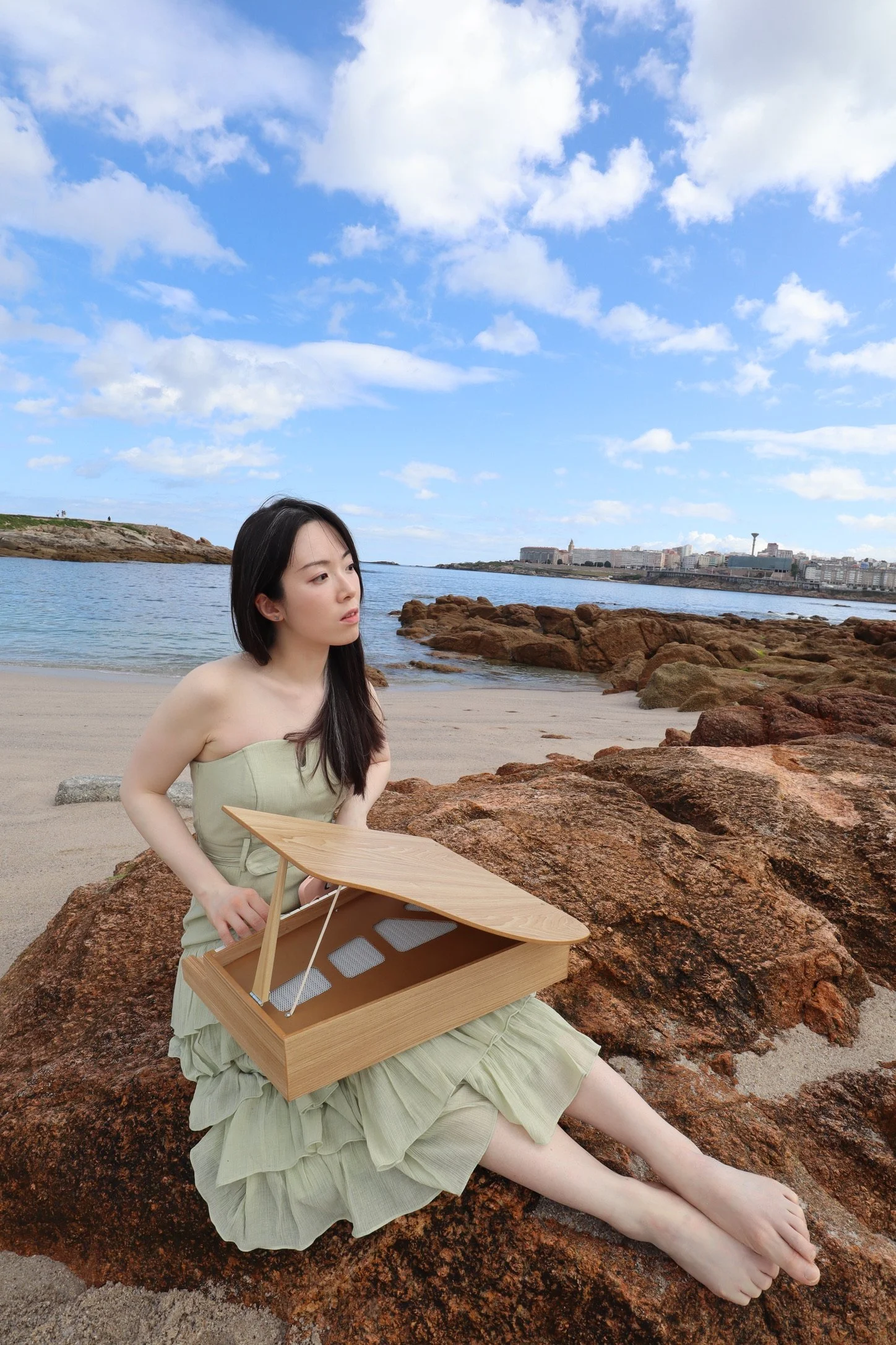 Jessica Wu, a young woman, sitting on rocks at a beach with a small piano, looking thoughtfully at the ocean under a partly cloudy sky.