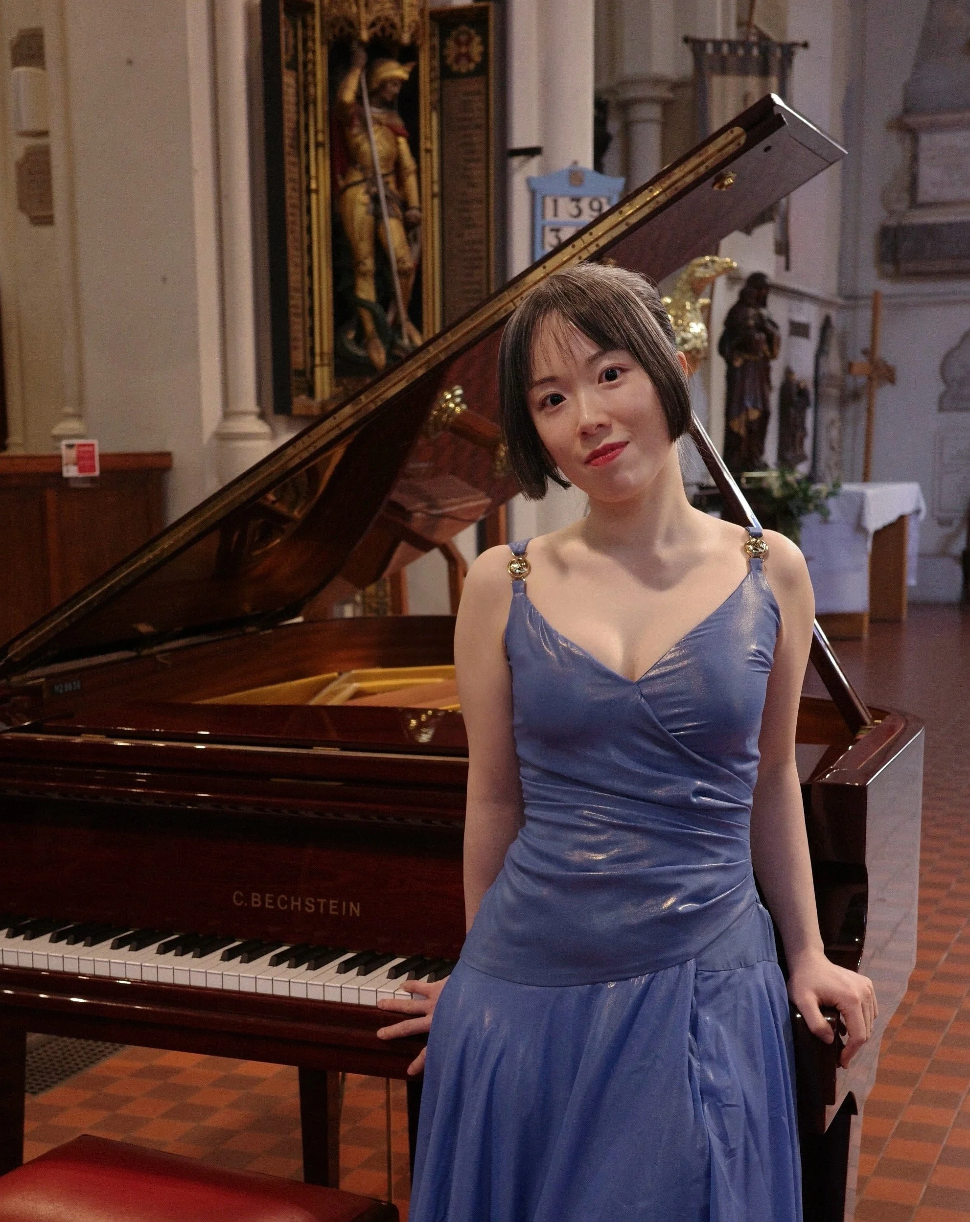 Jessica Wu, a young woman, in a blue dress standing next to a grand piano in a church or cathedral interior.