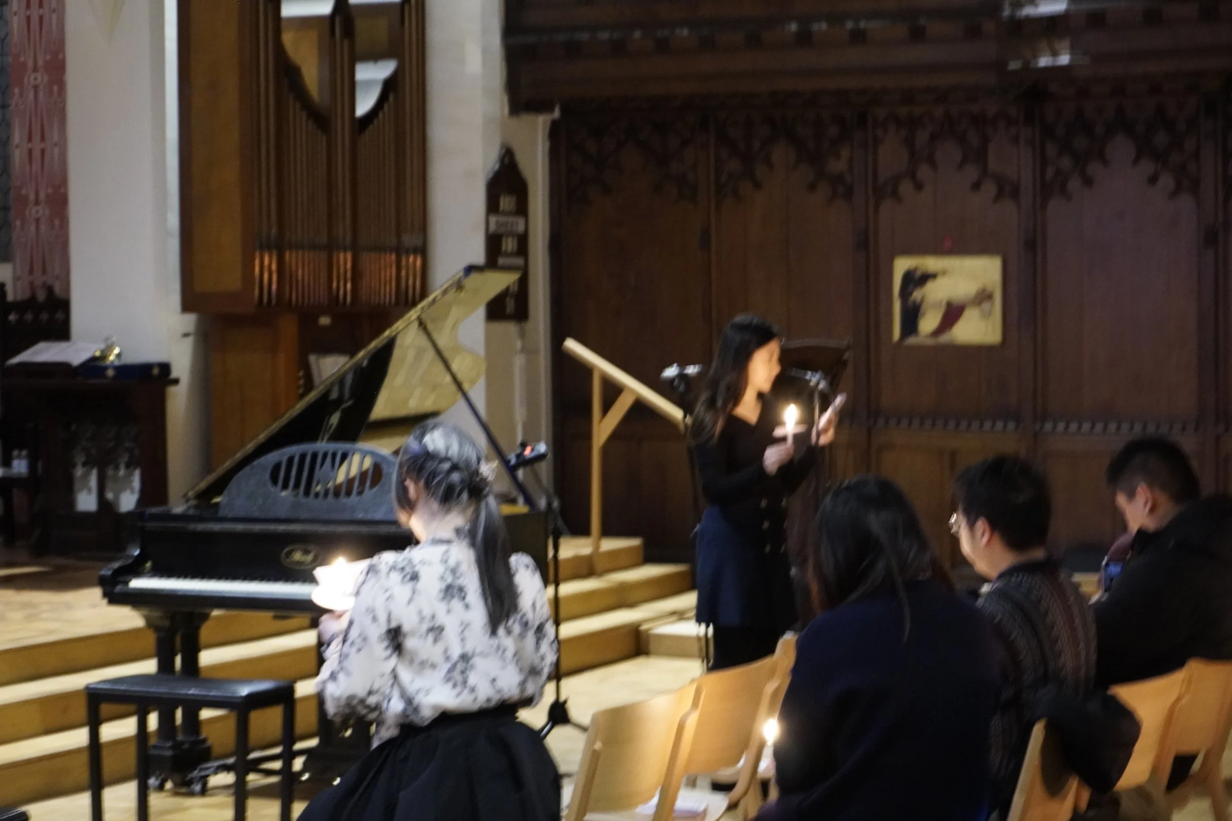 A woman stands on stage holding a candle and reading from a piece of paper, with a grand piano in the background and an audience seated watching.