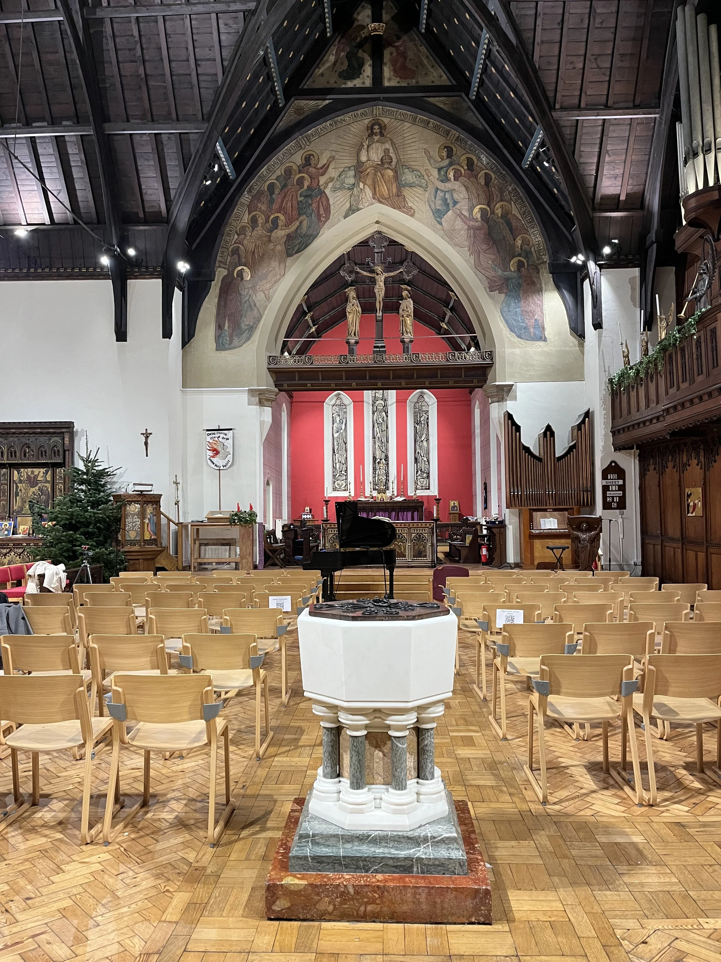Interior view of a church altar with a baptismal font in the foreground, rows of chairs, a Christmas tree, and religious artwork and statues.