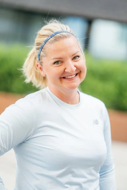 En smilende kvinne med blond hår og hvit treningstøy, ute i et grønt område.