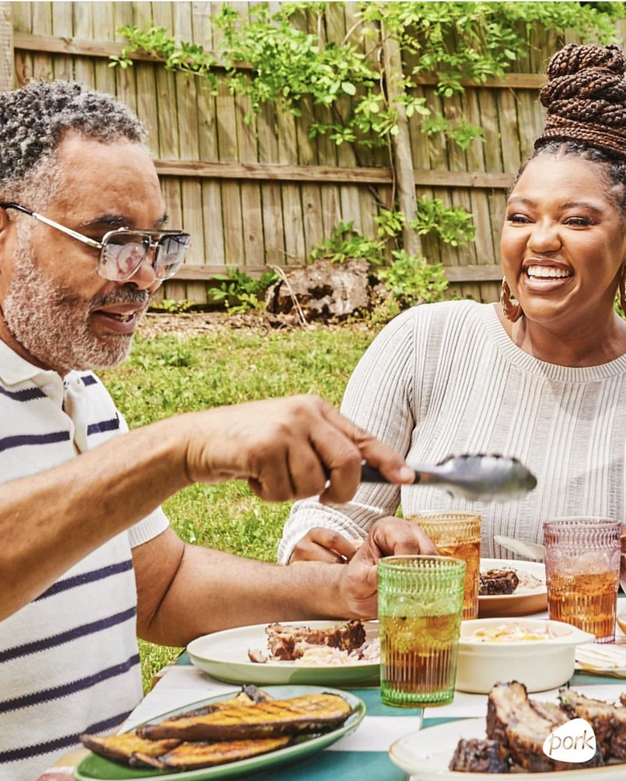 Two adults laughing and enjoying a meal outdoors at a backyard table.