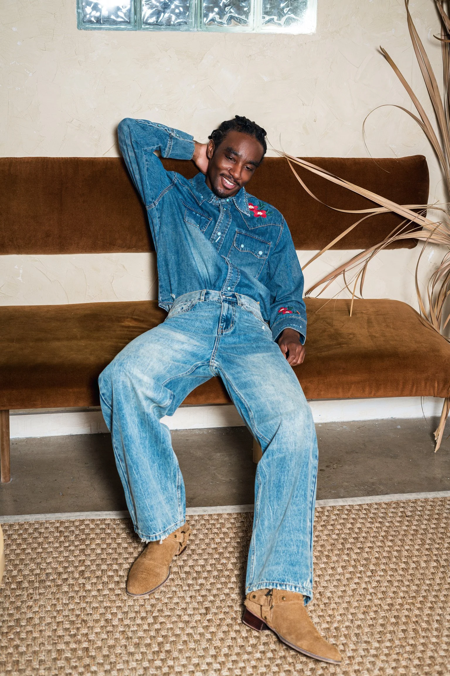 A man with dreadlocks smiling, sitting on a brown velvet bench, wearing a denim jacket with embroidery, light blue jeans, and tan suede boots, indoors with a beige textured wall, window, and large dried plant.