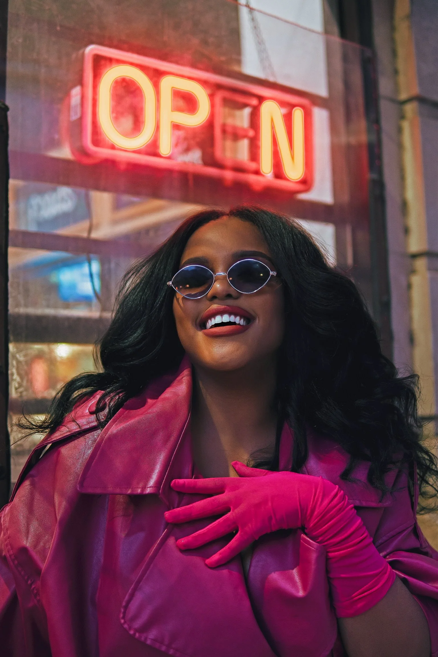 A woman in a pink jacket and gloves wearing sunglasses, smiling in front of a neon 'OPEN' sign at night.