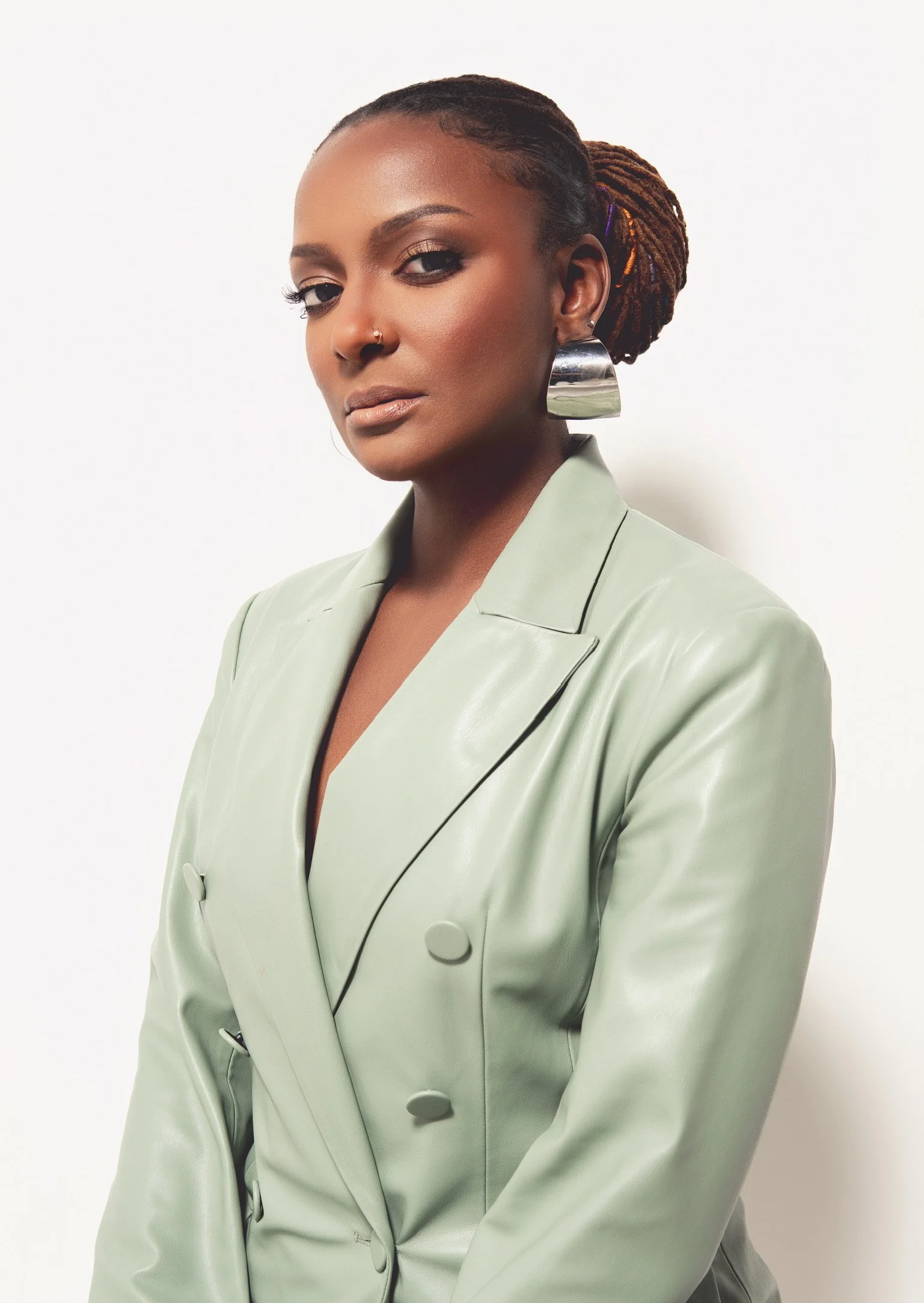 Portrait of a confident woman with dreadlocks wearing large silver earrings, makeup, and a light green blazer, standing against a plain white background.