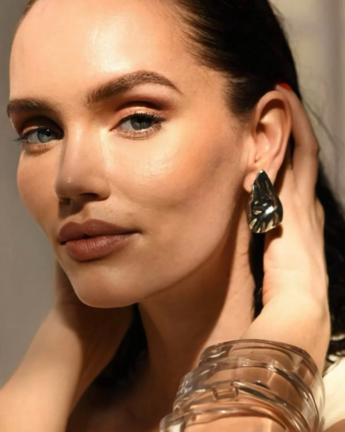 A close-up of a woman with fair skin, blue eyes, and dark hair, wearing makeup and large black and beige earrings, holding her hand near her face with bracelets on her wrist.