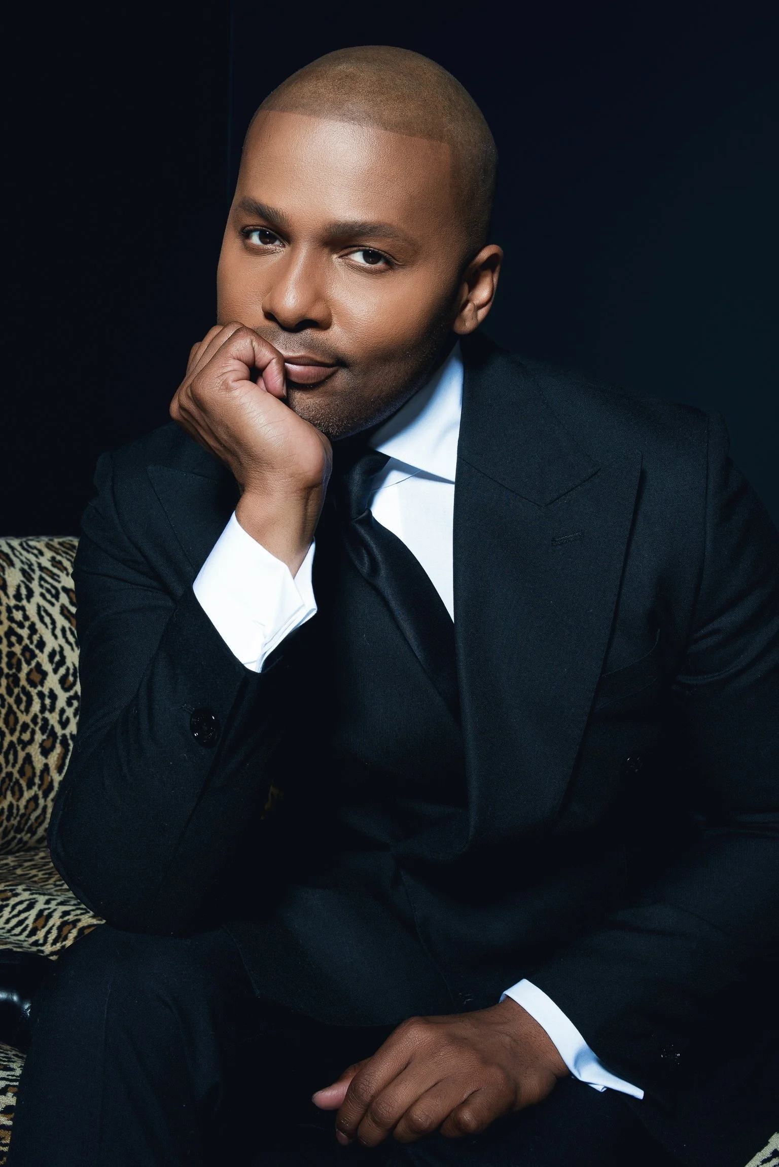 A man dressed in a black tuxedo with a white shirt sits on a leopard print chair, resting his chin on his hand, against a dark background.