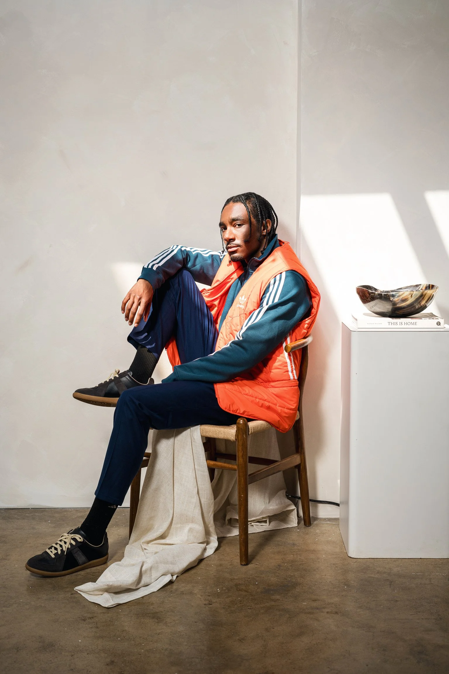 A man with braided hair sitting on a wooden chair in a minimalist room, wearing a blue tracksuit and an orange puffer vest, with an art piece on a white pedestal beside him.