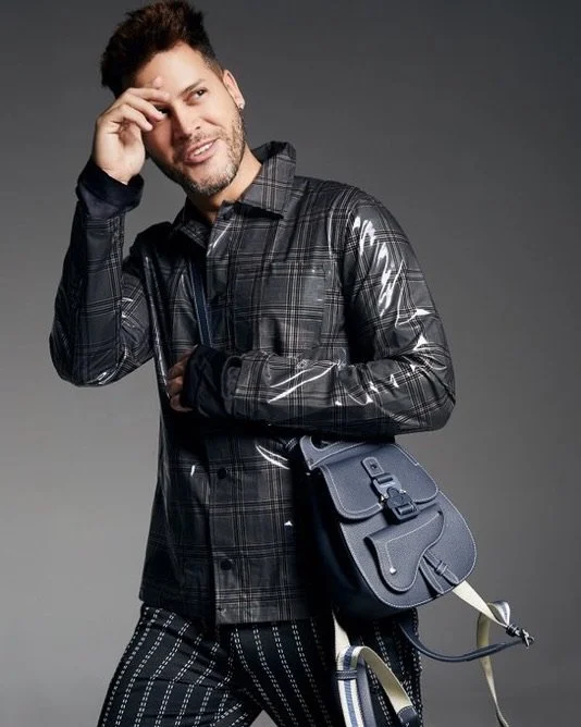 Man wearing a black plaid jacket with shiny accents, holding a gray bag with a strap, standing against a gray background.