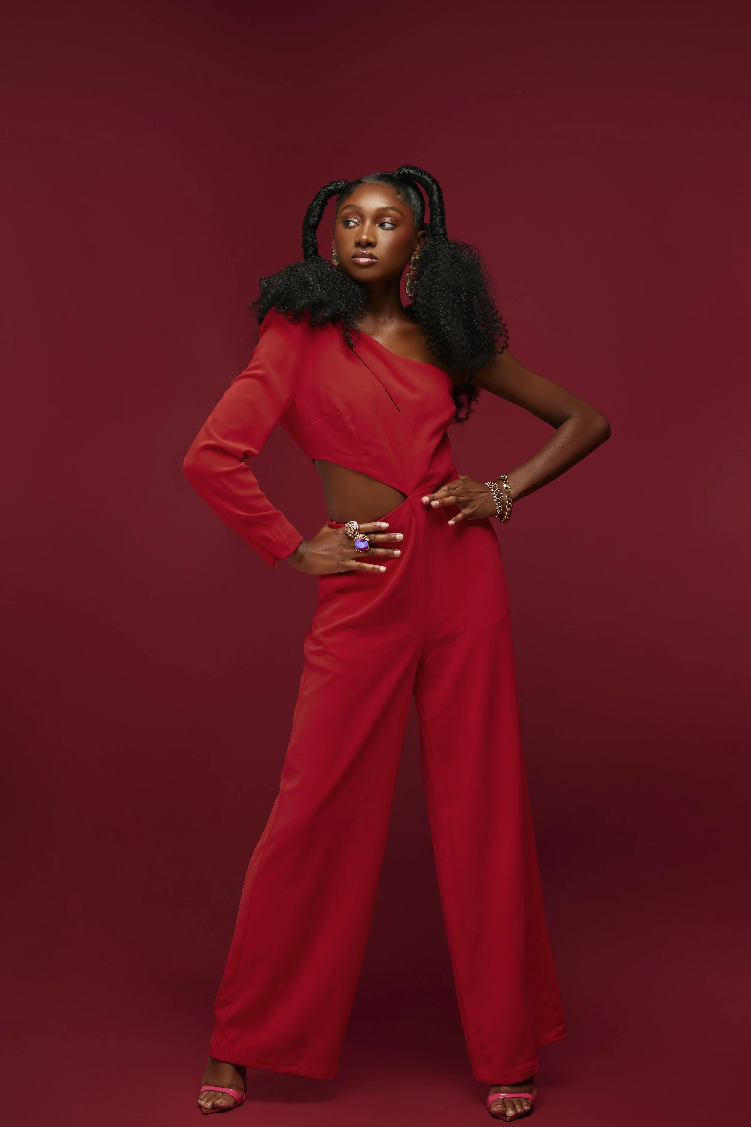 Woman in red jumpsuit with cut-out, standing against a red background, wearing jewelry and pink heels.