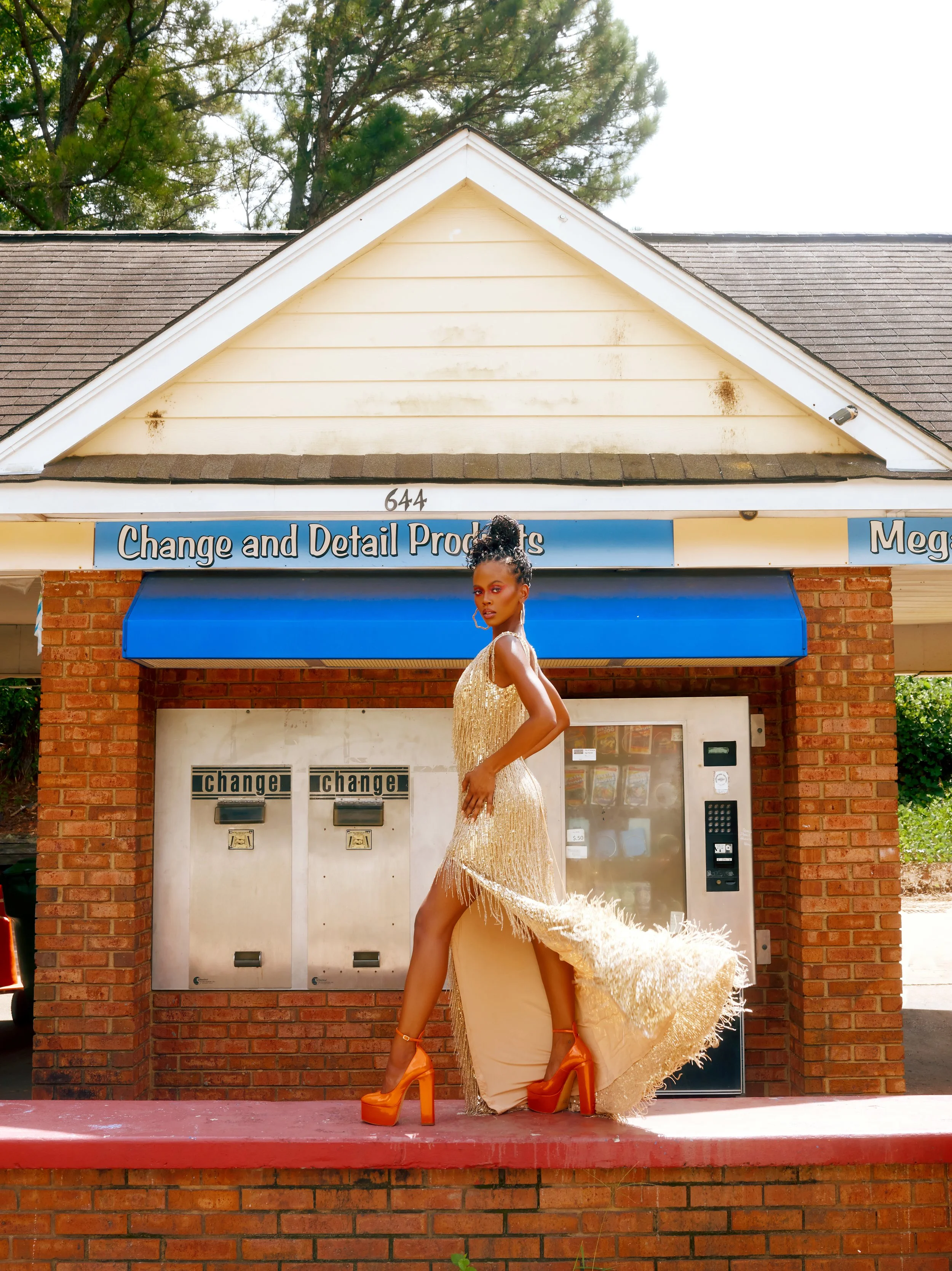 A woman in a gold, fringed dress and orange high heels stands in front of a vending machine outside a brick building with a blue awning, labeled 'Change and Detail Products.'