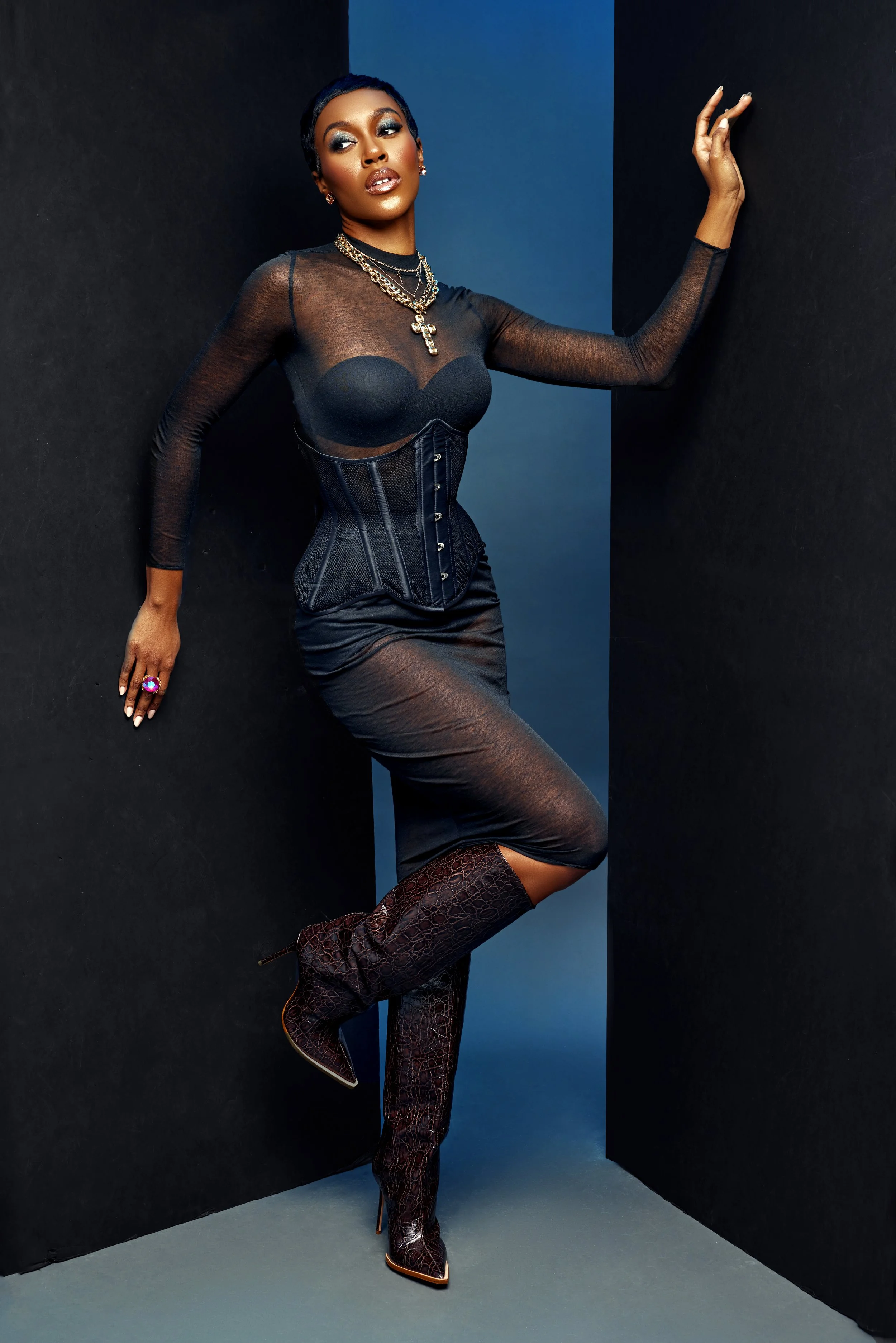A woman dressed in a sheer black outfit, layered necklaces, and high-heeled knee-high boots, posed against black and blue background.