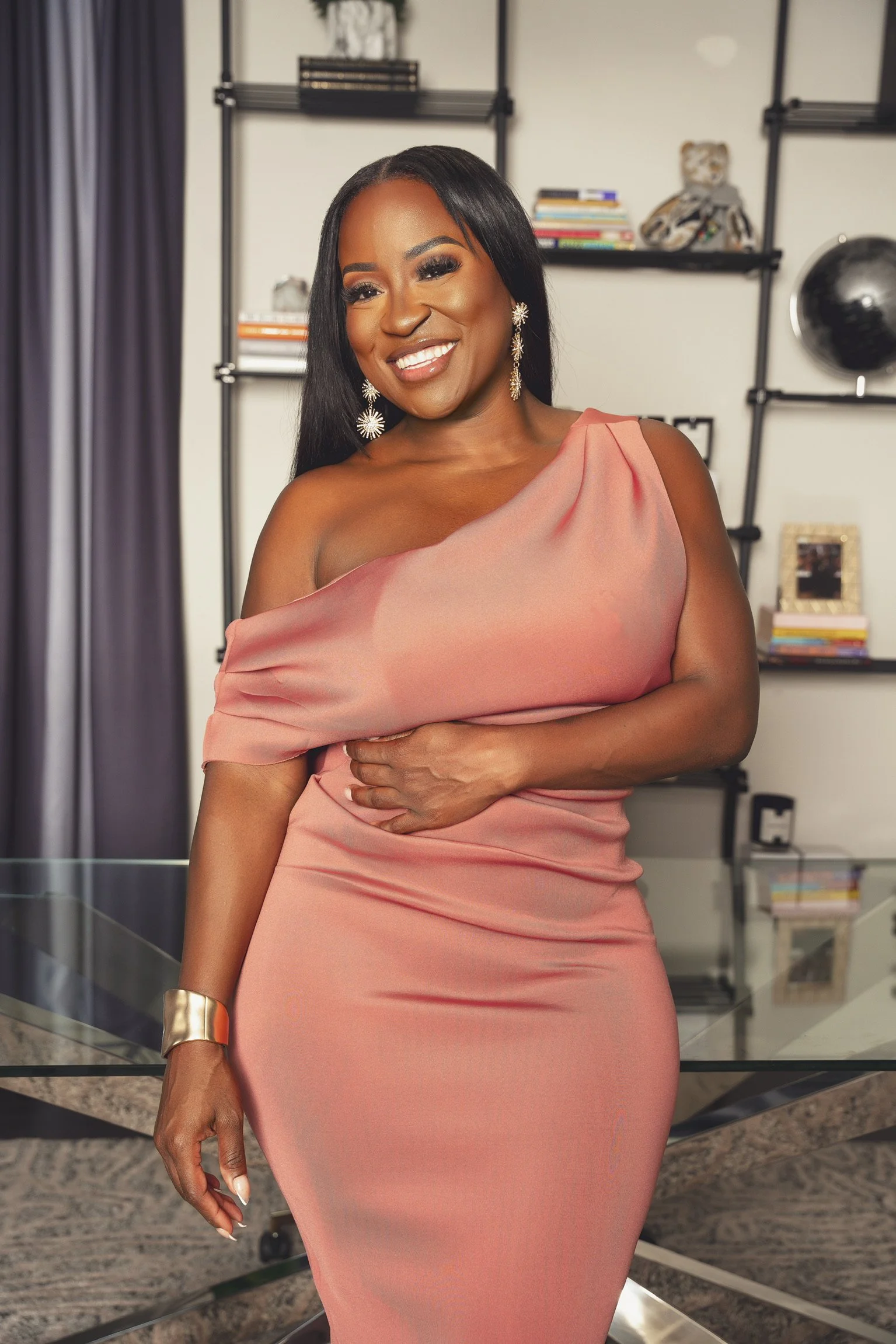 A woman in a pink off-shoulder dress standing in a modern room with shelves, books, and decorative items, smiling at the camera.