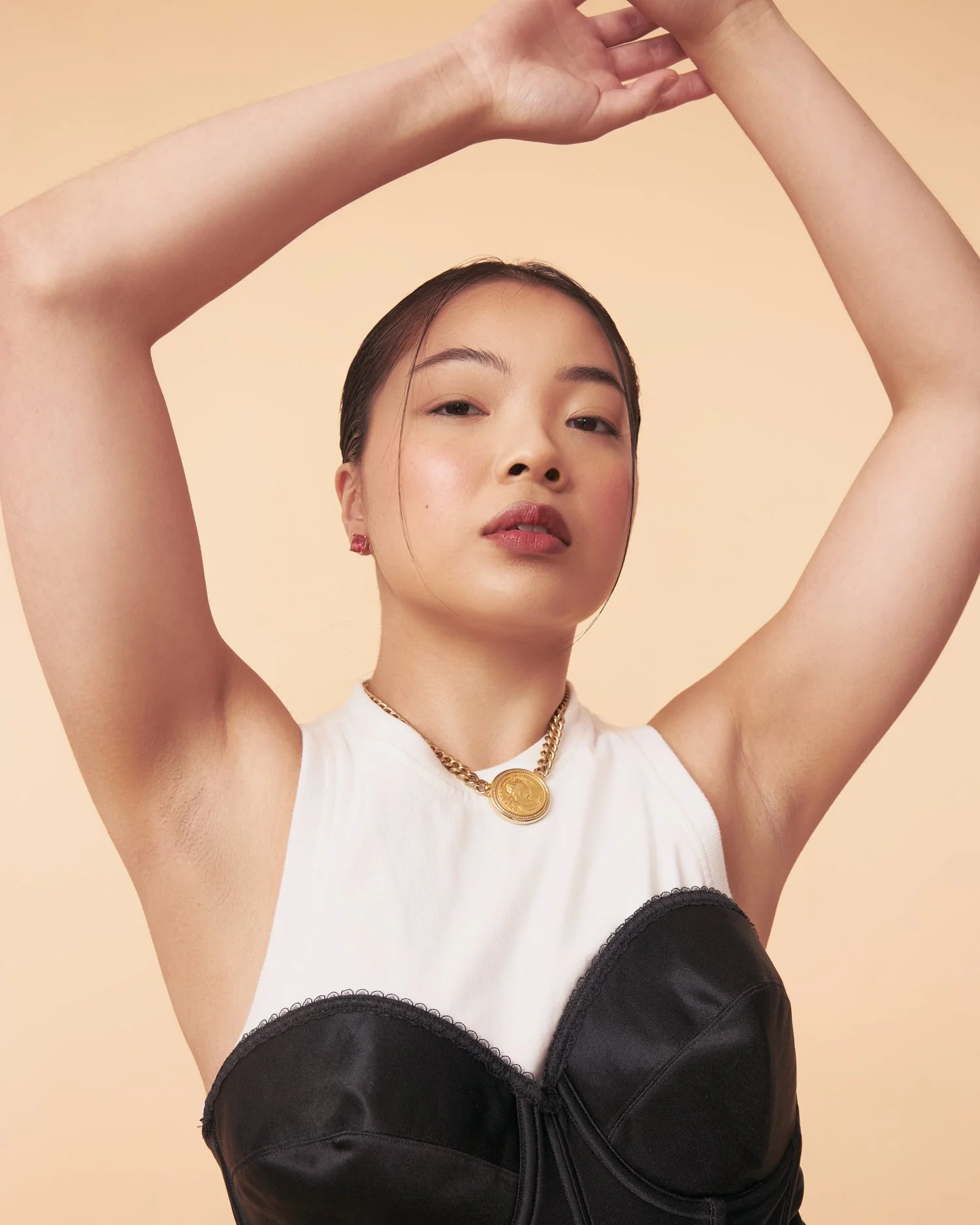 A woman with eyebrows raised and red lipstick poses with her arms raised above her head, wearing a white sleeveless top with black bustier and gold jewelry, against a beige background.