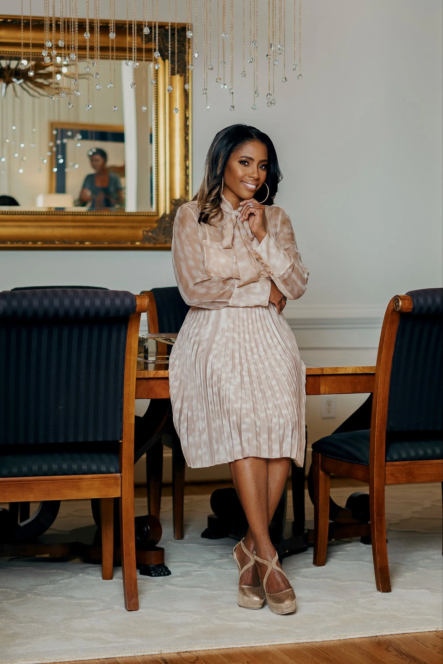 A woman in a beige patterned outfit and heels standing in a dining room with chairs and a mirror.