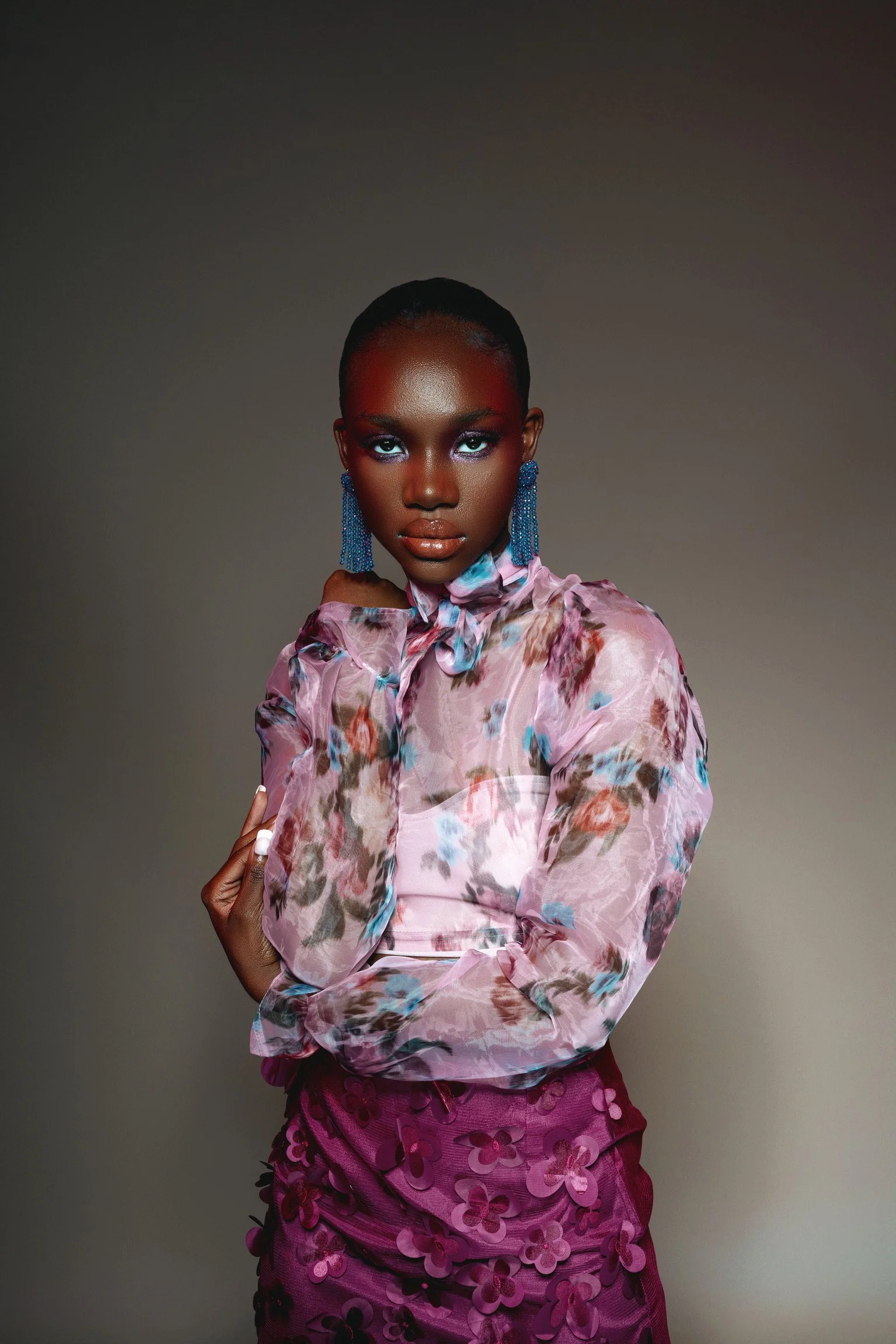 A confident woman with short hair, wearing bright blue earrings, a floral pink blouse, and a purple skirt with 3D flower details, standing against a dark gradient background.