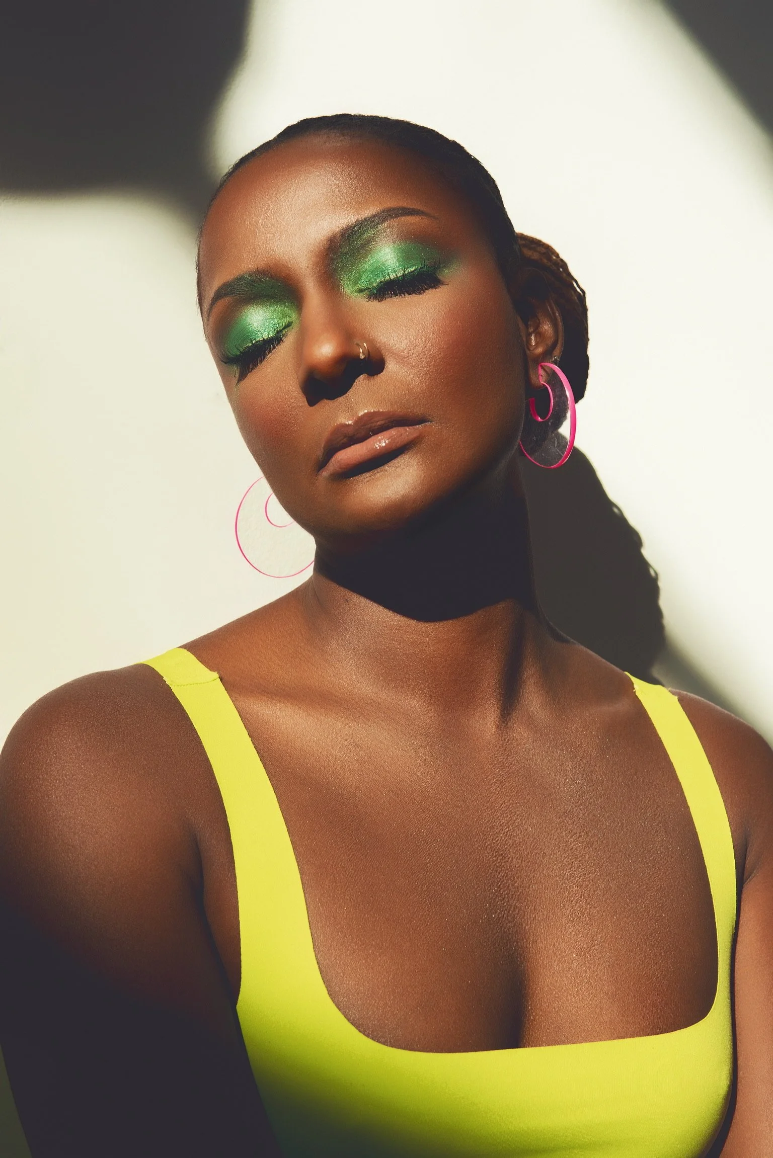 A woman with green eyeshadow, closed eyes, and pink hoop earrings, wearing a yellow top, with her face illuminated and shadowed.