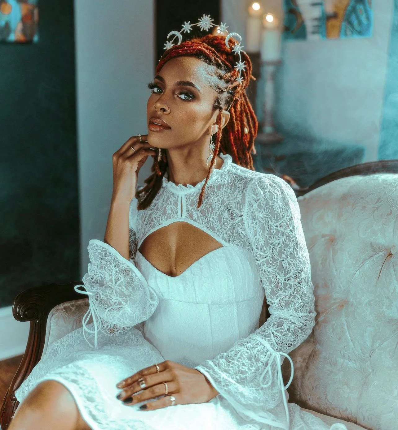 Woman with red dreadlocks wearing white lace dress and jewelry, sitting on a vintage sofa in a decorated room.