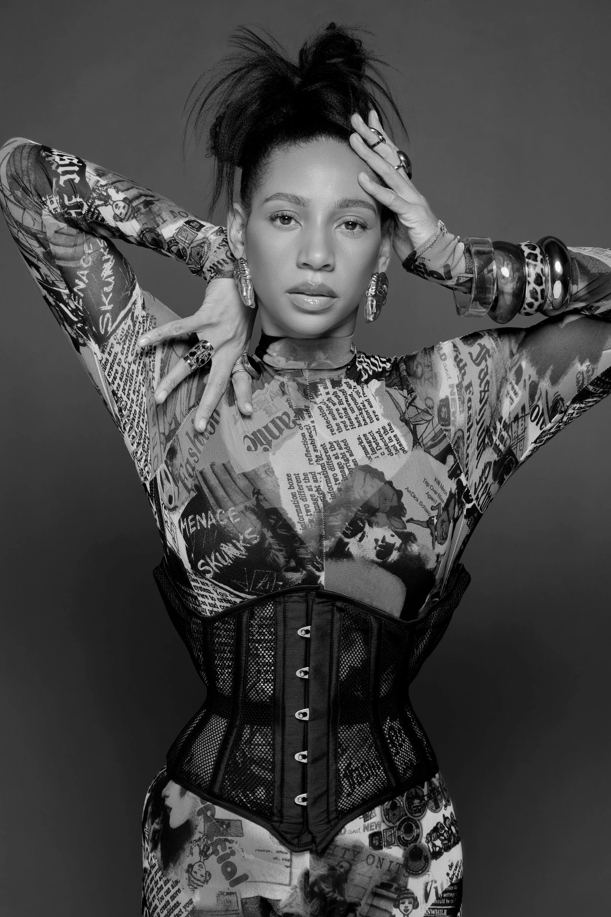 Black and white portrait of a woman with styled hair, wearing a newspaper print top, a waist corset, and multiple large bracelets and rings, looking directly at the camera with hands near her head.