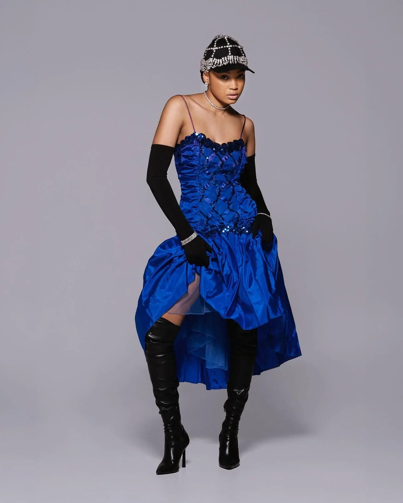 A woman in a royal blue dress with black gloves, thigh-high black boots, and accessorized with jewelry and a hat, standing against a plain background.