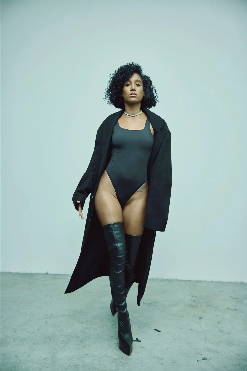 A woman standing against a plain background, wearing a black bodysuit, thigh-high black boots, a black coat draped over her shoulders, a pearl necklace, and small earrings, with curly hair and a confident expression.