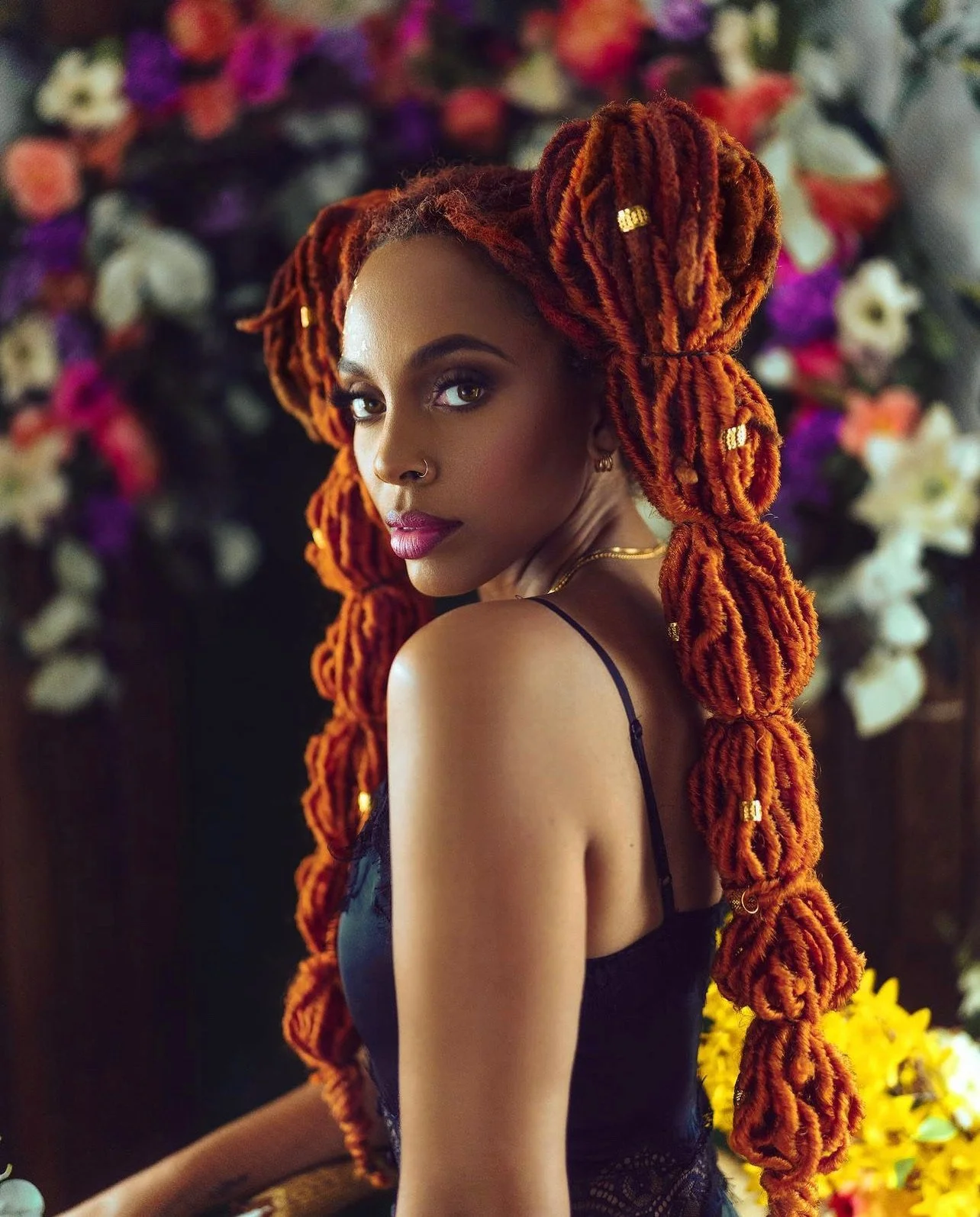 A woman with vibrant orange dreadlocks and a nose ring, wearing a black spaghetti strap top, posing against a colorful floral backdrop.