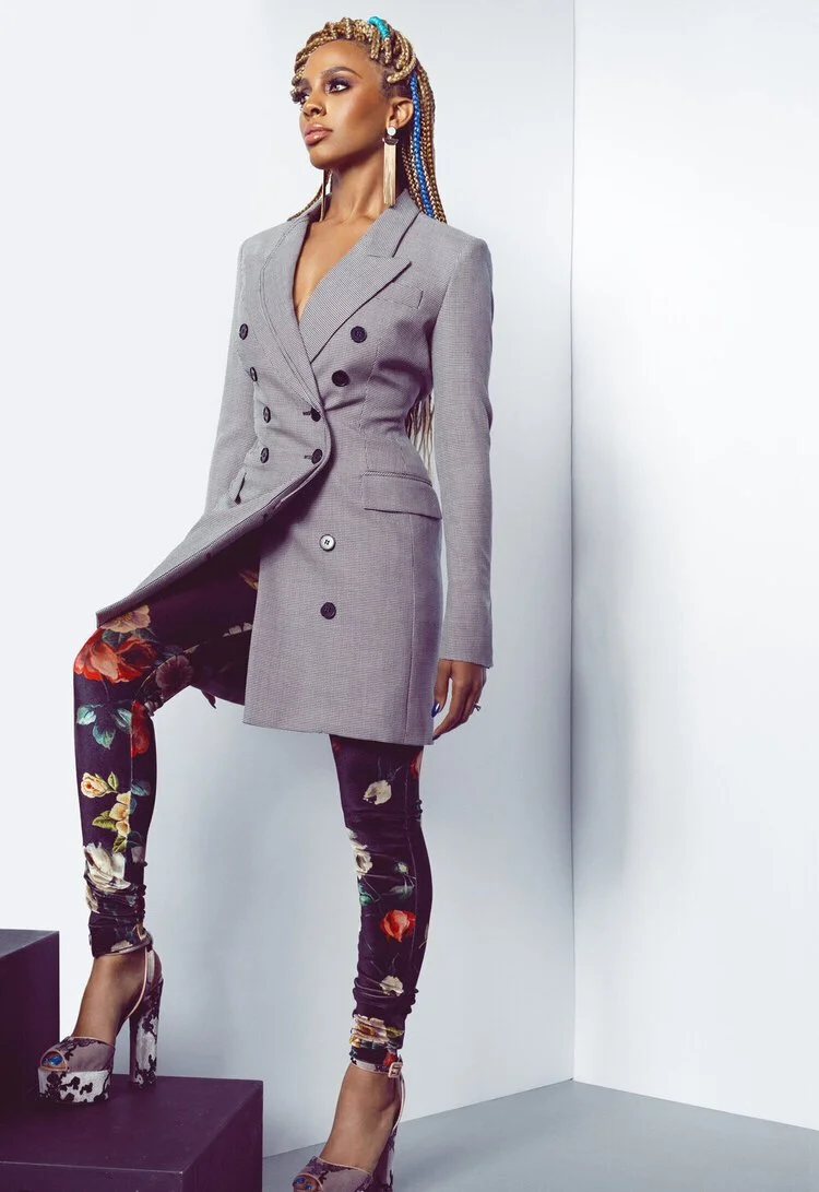 A woman with long braided hair wearing a gray double-breasted blazer dress, floral skinny jeans, and high heels, standing against a plain white wall.