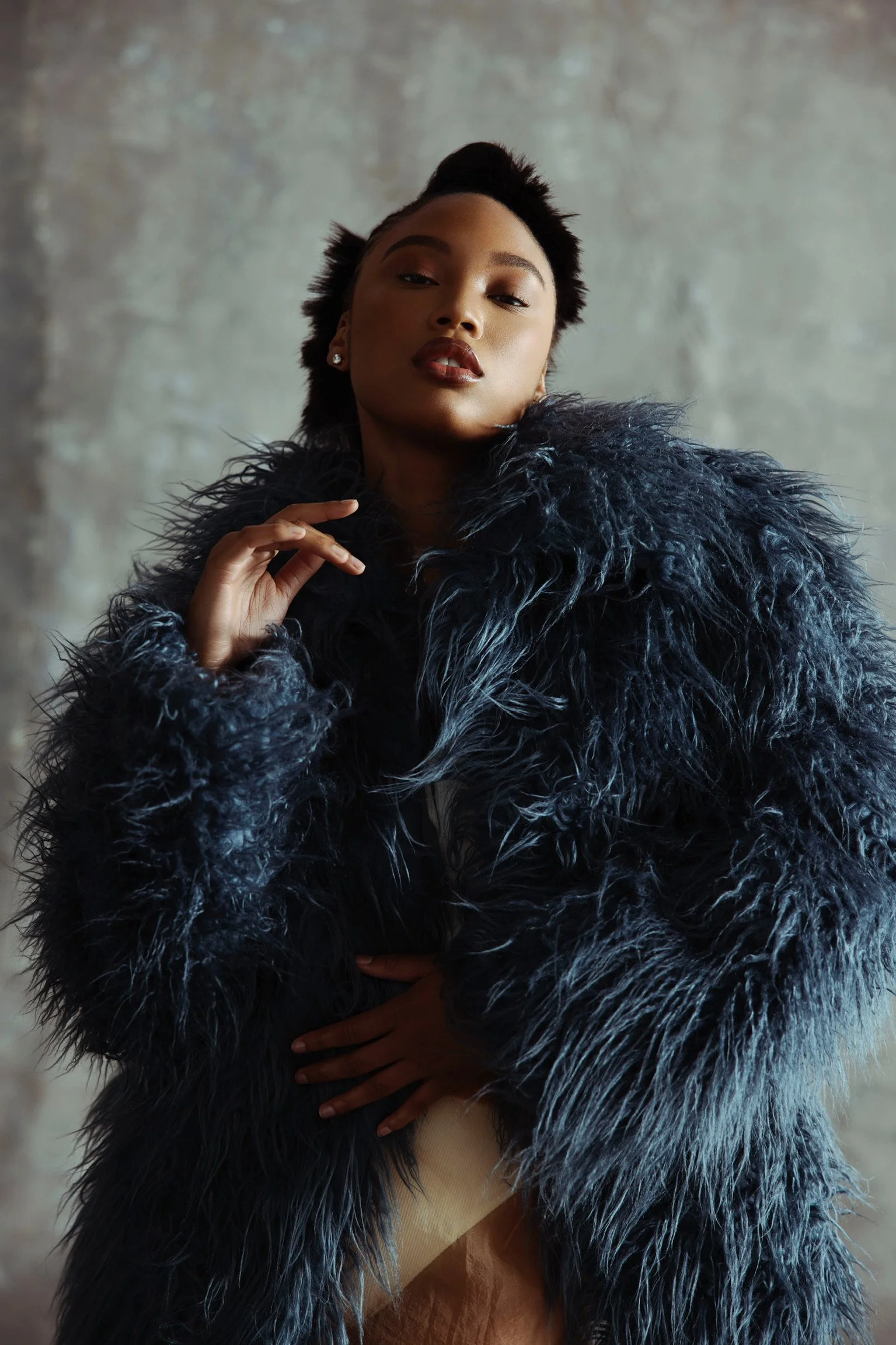 A woman wearing a dark, textured faux fur coat poses confidently against a neutral background.