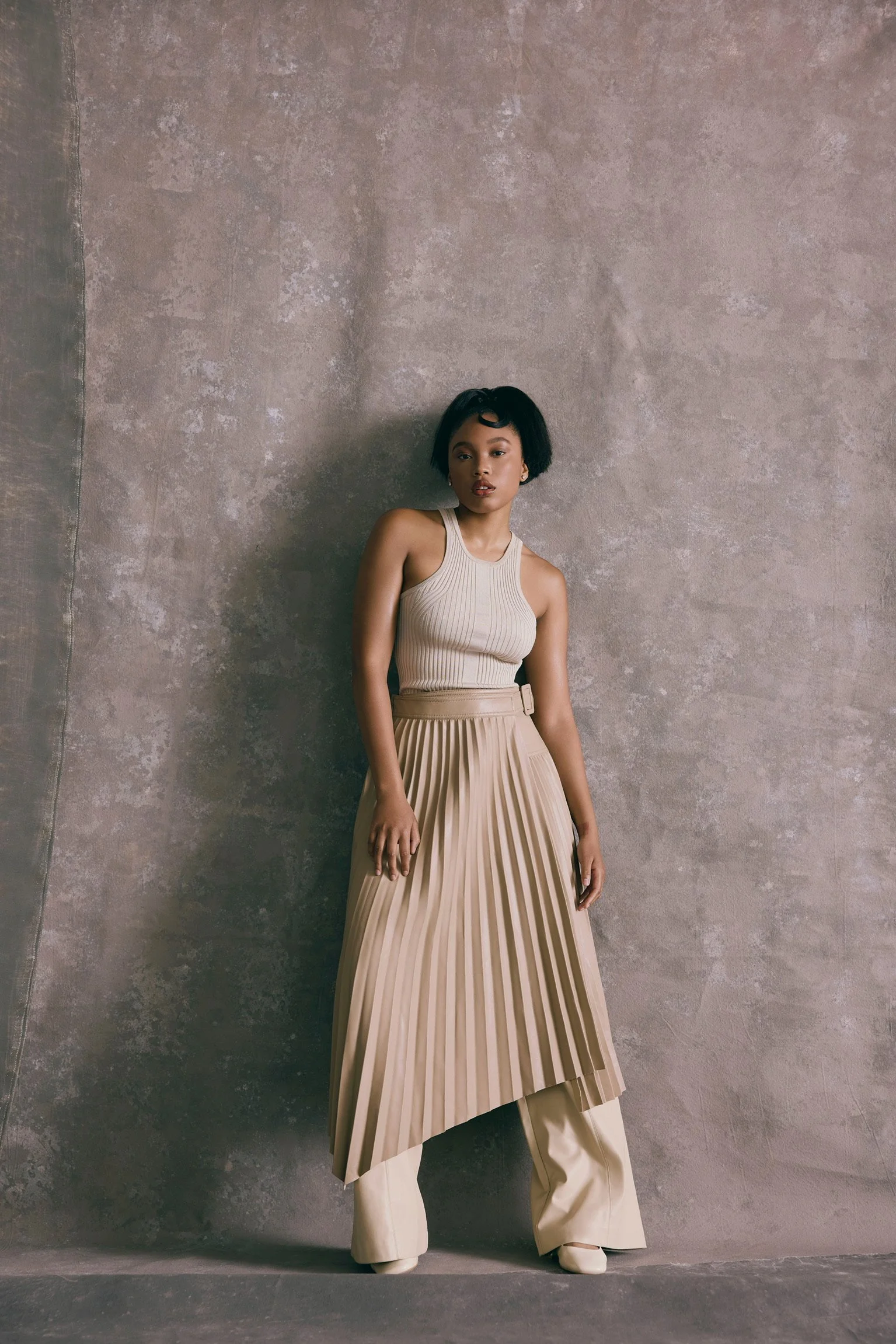 Woman in beige pleated skirt and tank top standing against a textured wall.
