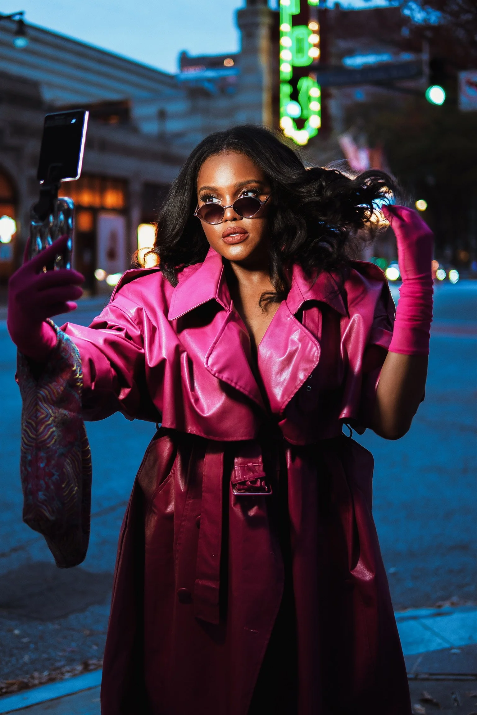 A woman with dark hair and sunglasses taking a selfie with her phone on a city street at dusk. She is wearing a shiny pink trench coat and pink gloves.