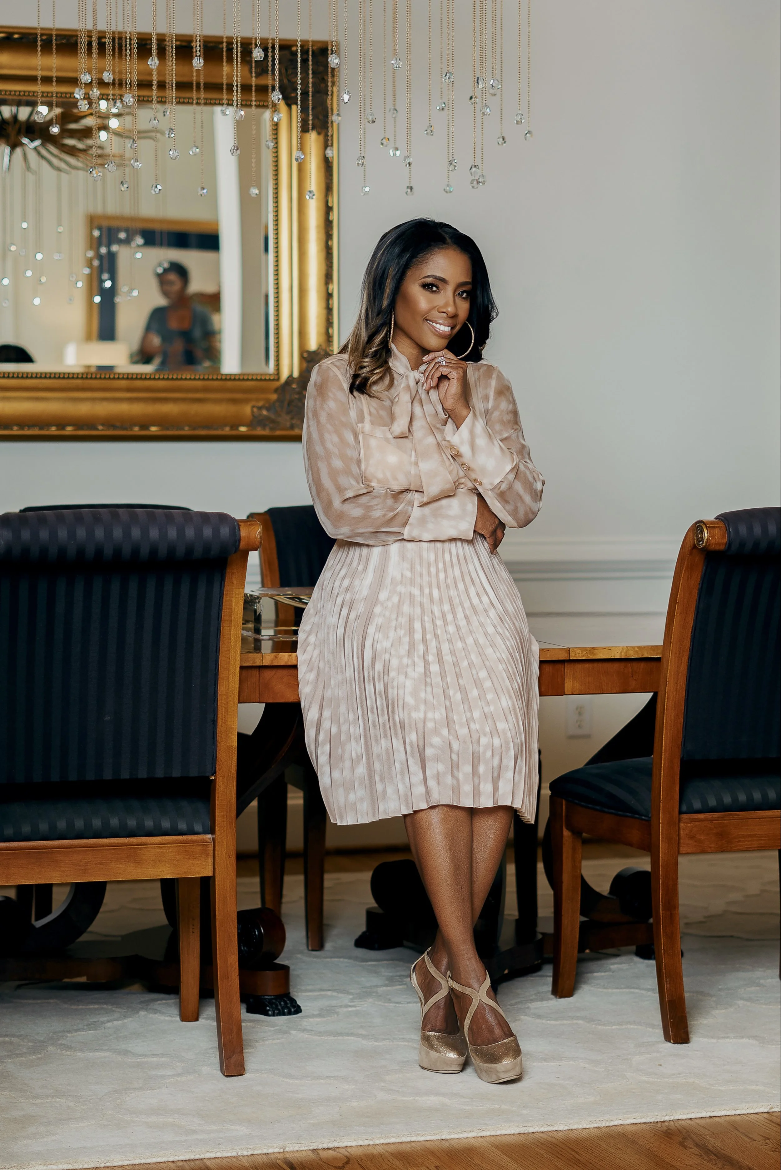 A woman dressed in a beige and pink outfit, sitting on a dining chair in a room decorated with a large mirror, chandelier, and a dining table, smiling and posing for the camera.