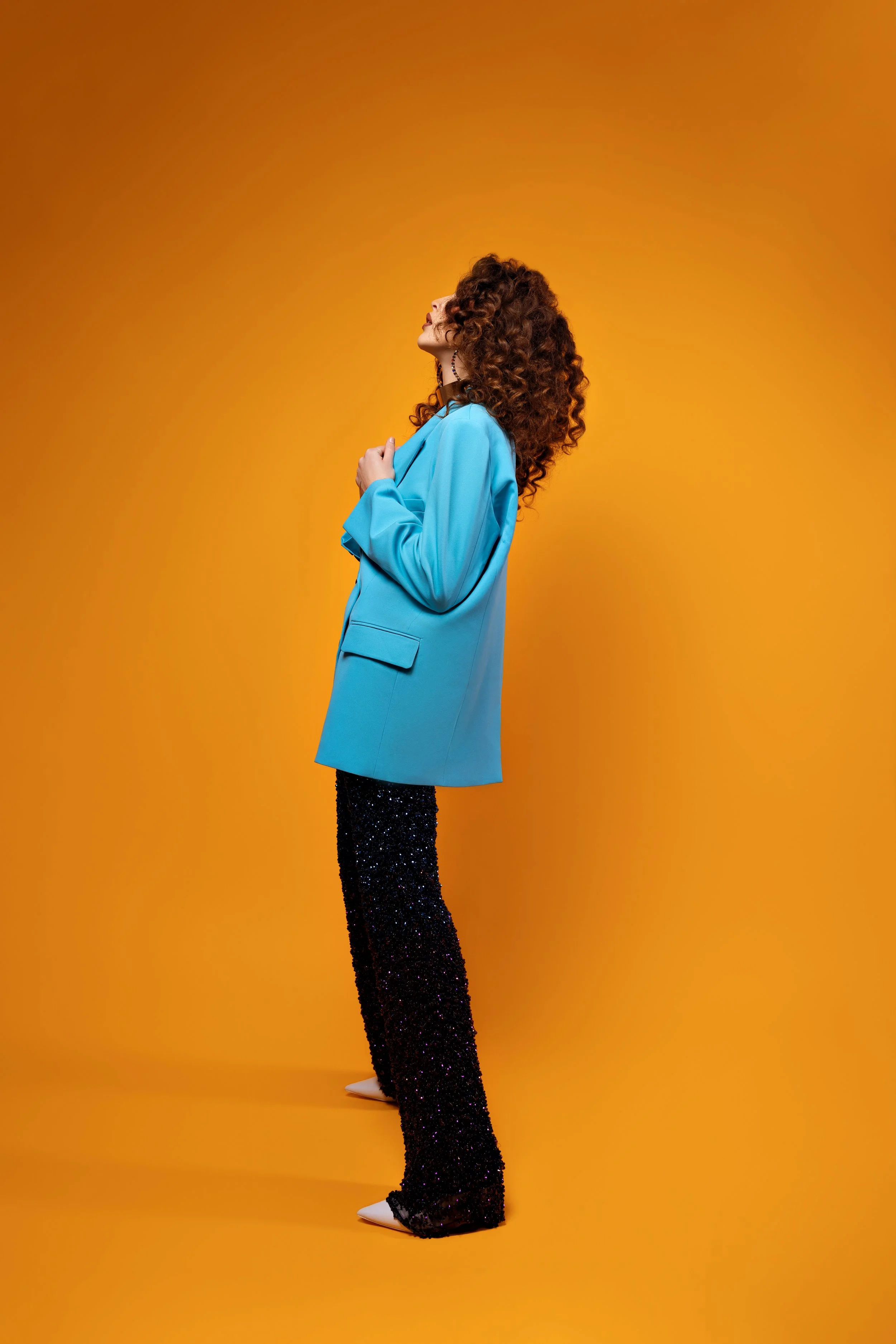 A woman with curly red hair wearing a bright blue blazer and black sparkly pants, standing against an orange background.
