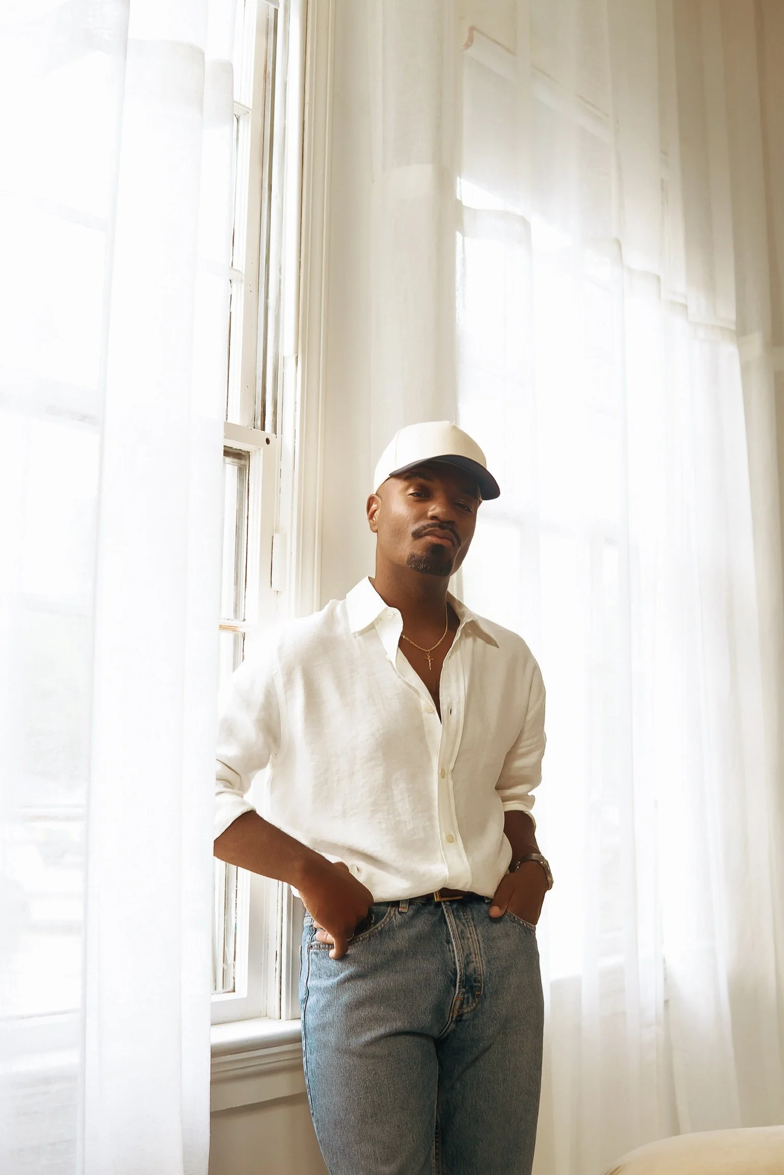 A man standing indoors near a window with sheer white curtains, wearing a white shirt, blue jeans, a white cap, and accessories.